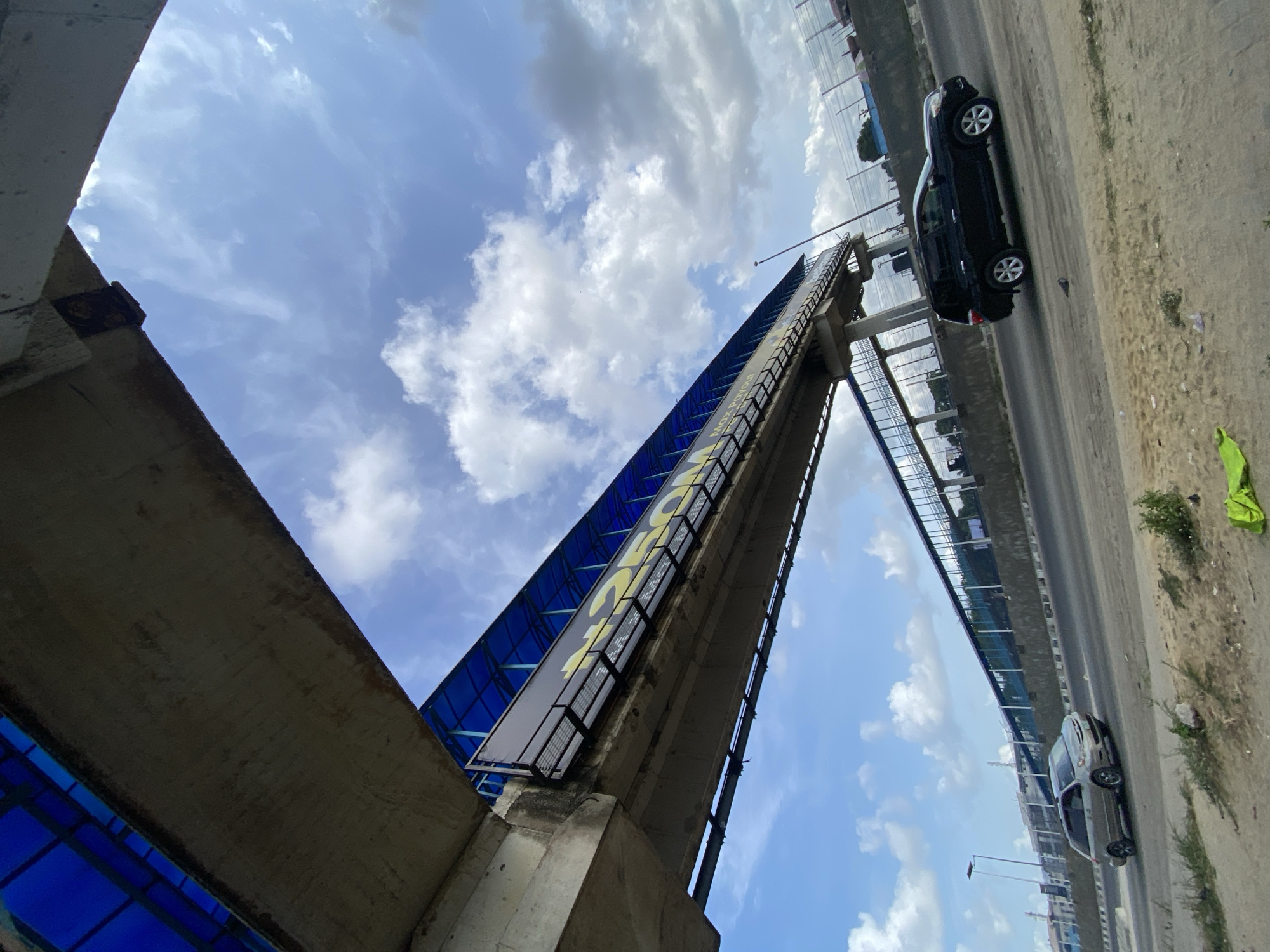 The image shows a view of a pedestrian bridge spanning a highway in Lekki, Nigeria. The bridge has a blue-tinted roof and signage. Two vehicles, a black SUV and a silver sedan, are visible on the road below. The sky is partly cloudy. Vegetation is seen on the side of the road. The perspective is from below, looking upwards at an angle.
