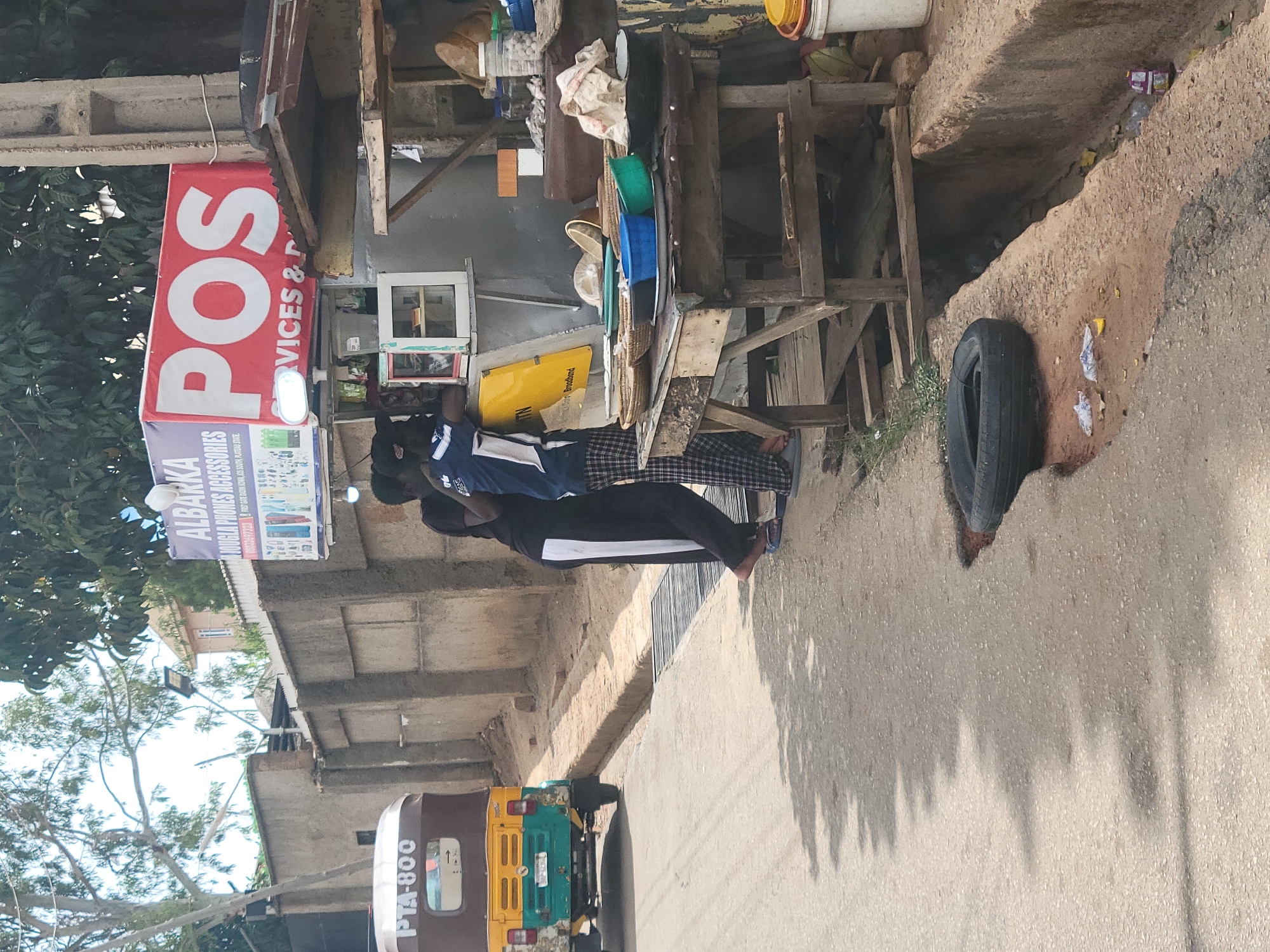 Here's a concise description of the image:
The image shows a street scene, presumably in the Jos area of Nigeria, judging by the signage. A small roadside stall labeled "ALBARKA JOUGLA PHONES ACCESSORIES" and "POS SERVICES &…" is visible. Two men are standing in front of the stall. A three-wheeled vehicle (Tuk-tuk or Keke Napep) with the number "PTA-800" is parked on the road. A used tire rests against the curb. The environment appears to be a mixed residential/commercial area with trees and concrete buildings nearby.