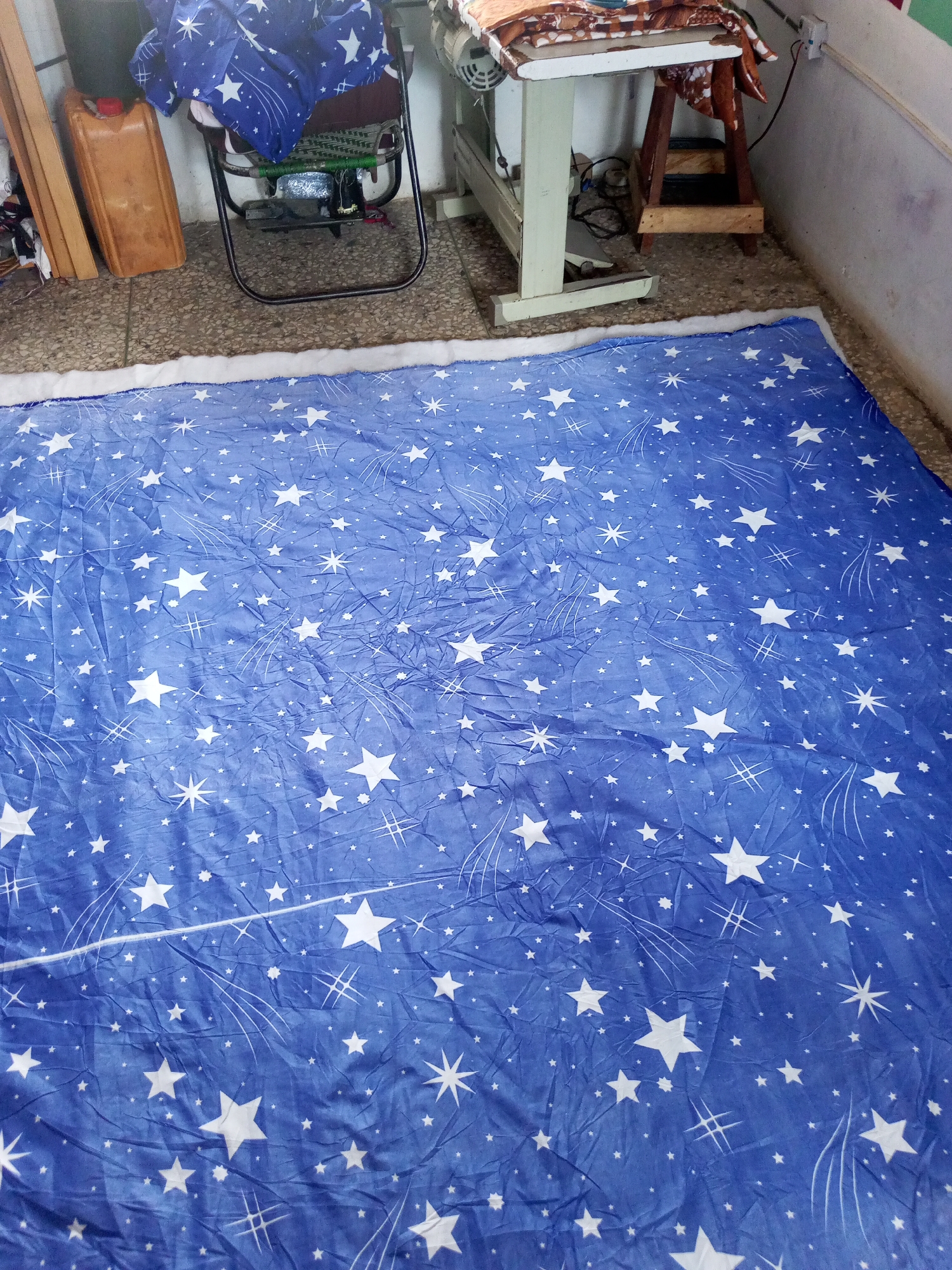An overhead, wide shot captures a large, blue blanket patterned with white stars and meteor streaks, laid out on a textured, tiled floor. The blanket is wrinkled, suggesting it's made of a soft fabric. A white, fluffy border runs along the top edge of the blanket. In the background, to the left, a brown plastic container and wooden planks are visible. A metal folding chair, piled with folded blue and patterned fabric, stands next to them. To the right, a white sewing machine with a wooden base and a wooden leg is positioned against a light-colored wall. The environment appears to be an indoor workshop or storage area, with the floor showing signs of wear.