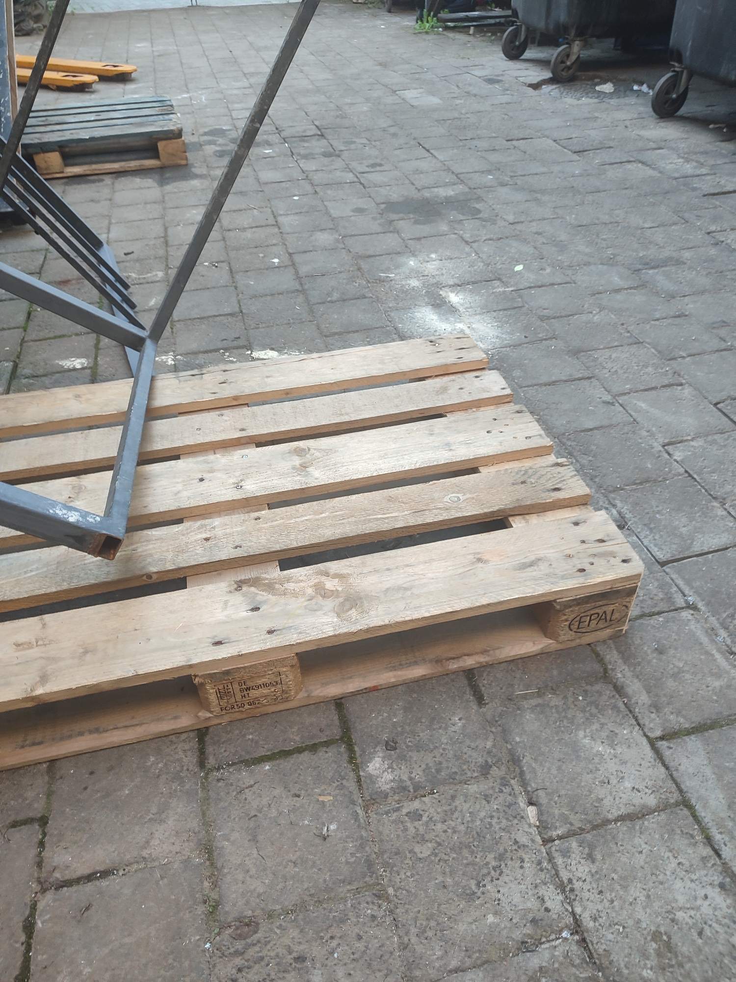 The image shows a close-up of a wooden pallet resting on a paved surface, likely in Chernivtsi, Ukraine. A portion of a metal structure is also visible, leaning against the pallet. The paved area appears to be made of interlocking concrete blocks, and there are some scattered debris and white powder on the ground. In the background, large black trash bins with wheels are partially visible. The scene is outdoors, and the lighting suggests it might be daytime. There are no people or animals present in the frame. The pallet has markings indicating it is an EPAL pallet, and further stamps are visible on one of its blocks, including "DE BW4911053 HT" and "FCR50 052-5".