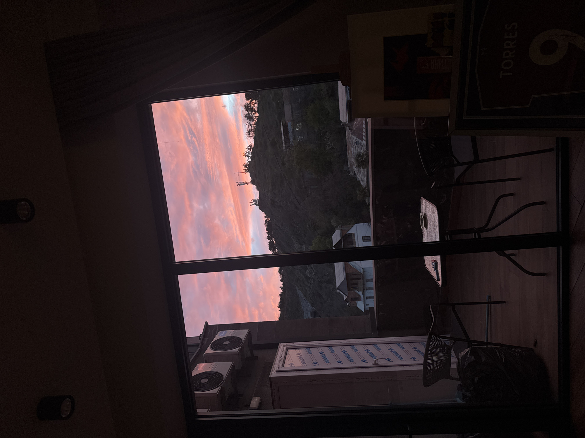 The image displays an indoor perspective looking out through a large window or glass door during either sunrise or sunset. The sky is a prominent feature, filled with dramatic pink, orange, and purple clouds, indicating the early morning or late evening. Beyond the window, a dark, tree-covered hillside or mountain dominates the mid-ground, with indistinct buildings and what appear to be communication towers visible on its slopes. Further in the distance, a few light-colored buildings can be seen.
Inside the dimly lit room, the immediate foreground on the left shows a wall with two dark wall-mounted light fixtures. On the right, there's a dark floor where two dark metal chairs with woven backs and a small table are arranged. One chair is pulled up to the table, and a dark bag is visible on the floor near another chair. Higher on the wall to the right, a framed poster is visible, showing the text "TORRES" and a large number, possibly "6" or "9." Another partial poster is below it.
On an outdoor ledge or balcony just outside the window, two white air conditioning units are mounted. Below them, a rectangular object covered in white protective sheeting, repeatedly displaying the word "panoline," suggests construction or new installation. No people are visible, and the scene is static, capturing a moment of natural beauty and an ordinary interior setting.