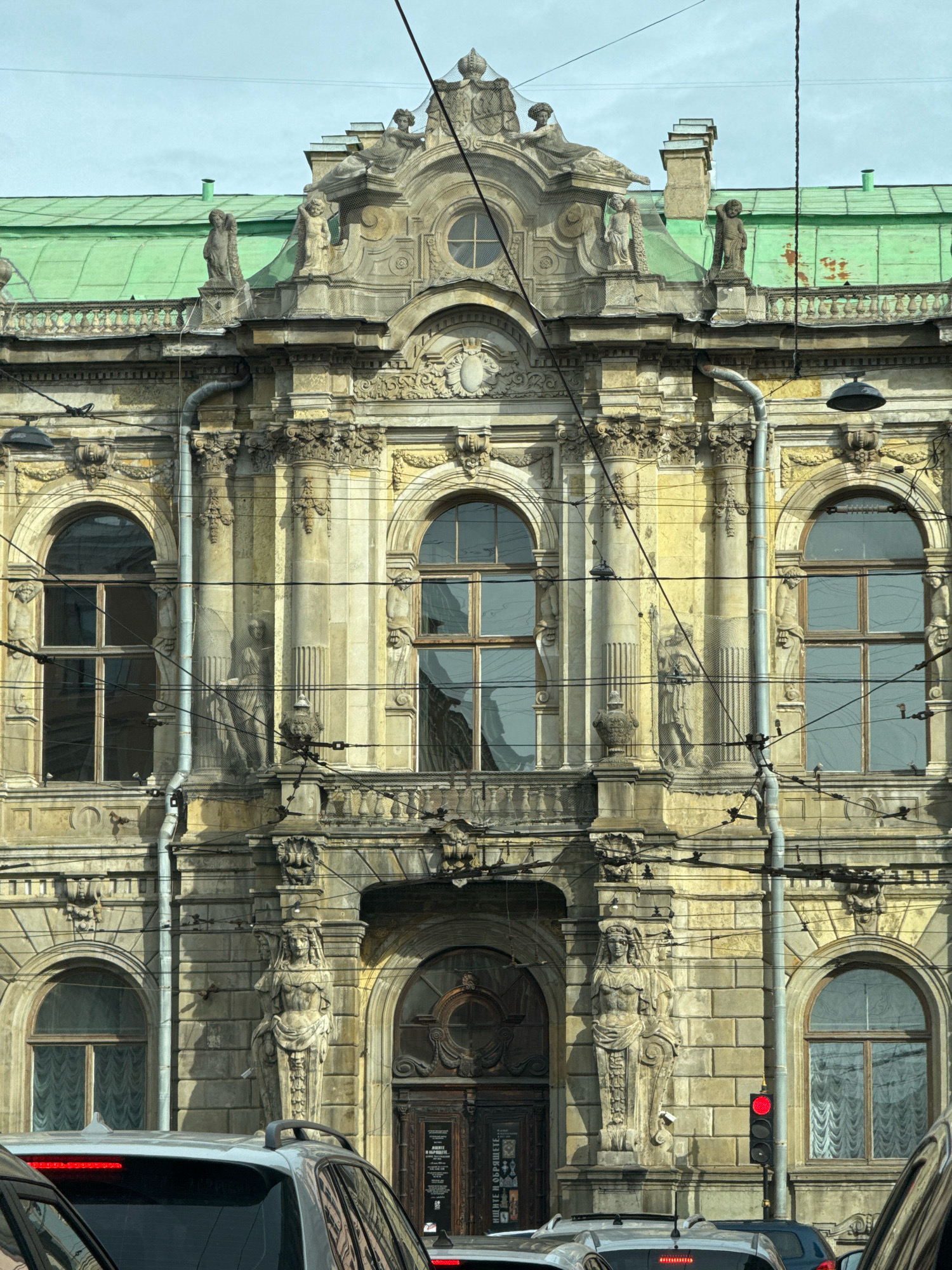 A close-up, eye-level, full shot captures the ornate facade of a historic building in Saint Petersburg, Russia, bathed in daylight. The building's weathered, pale yellow stone is adorned with classical architectural elements, including sculpted figures, columns, and elaborate carvings. Three arched windows with dark frames and reflective glass are visible on the upper level, each flanked by pilasters. Below these, a balcony with a decorative railing overlooks a grand entrance with a dark, arched wooden door. Two large, standing female sculptures flank the entrance, wearing draped clothing and elaborate headdresses.
The building's green, sloped roof is partially visible at the top, with some areas showing signs of age and rust. Numerous overhead wires crisscross the scene, obscuring parts of the facade and suggesting the presence of a street or thoroughfare. In the foreground, the tops of several vehicles are visible, including a silver car with its taillights illuminated, indicating they are stopped or moving slowly. A traffic light with a red signal is also present, further confirming the urban street setting. The overall impression is of an aged, grand architectural structure in a bustling city environment.