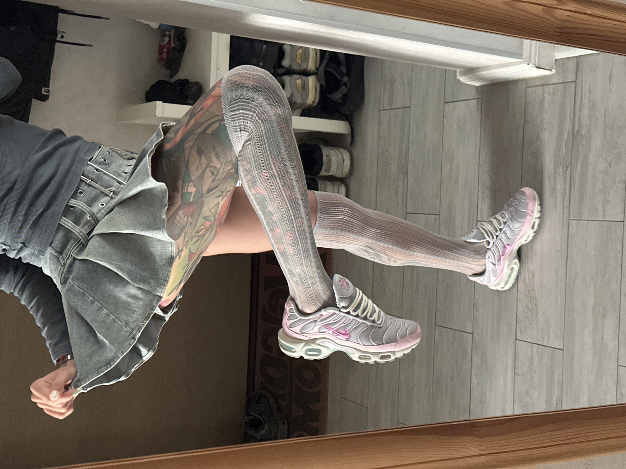 The image shows a person's legs reflected in a mirror. The person is wearing a grey denim skirt with a cinched waist, a long-sleeved grey top, and white patterned tights. They are also wearing grey and pink Nike sneakers. The person has tattoos on their legs. The background appears to be a room with tiled flooring and shelves filled with shoes. The reflection suggests the photo was taken indoors, likely in a personal space like a bedroom or dressing area, with a wooden frame visible at the bottom of the image. The overall mood is casual and stylish.