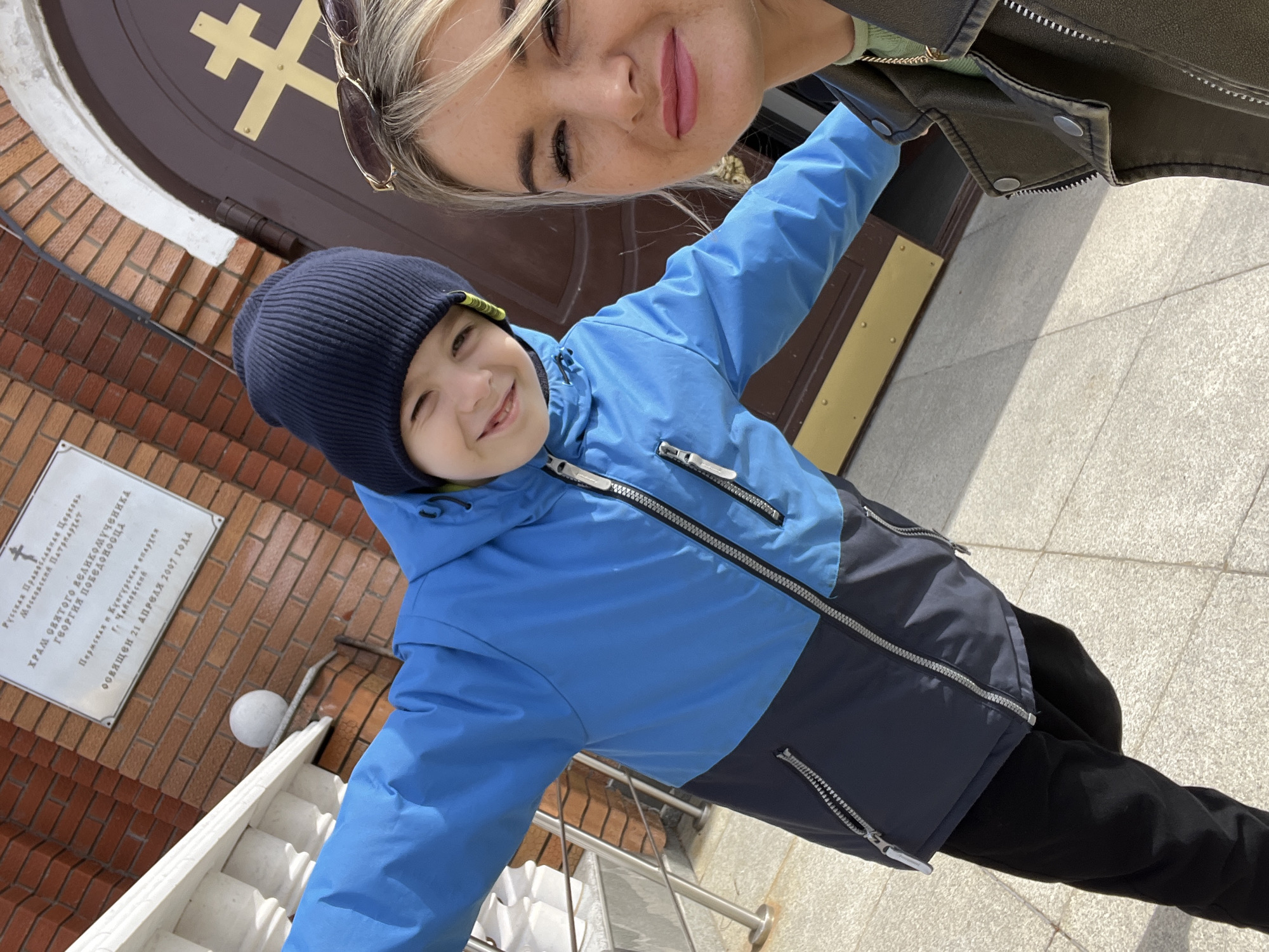 A woman and a young boy are depicted in a close-up selfie taken outdoors in bright daylight. The woman, with blonde hair and sunglasses pushed onto her head, is partially visible at the top of the frame, smiling with her eyes slightly closed. The boy, positioned below her, wears a navy blue beanie and a blue and dark blue two-tone jacket. He is smiling widely, looking directly at the camera with an open, joyful expression, and has his arms slightly spread.
They are standing in front of the entrance to a church building. The background features a reddish-brown brick wall and a dark brown door adorned with a prominent golden Orthodox cross. To the left of the door, a white rectangular plaque is mounted on the brickwork. White-painted stairs with a balustrade are visible on the left side, leading up towards the entrance. The ground is paved with light grey tiles or concrete, illuminated by sunlight which casts shadows.
The visible text on the white plaque, written in Russian, identifies the location as the "Church of the Holy Great Martyr George the Victorious" (ХРАМ СВЯТОГО ВЕЛИКОМУЧЕНИКА ГЕОРГИЯ ПОБЕДОНОСЦА) belonging to the "Russian Orthodox Church, Moscow Patriarchate" (Русская Православная Церковь, Московский Патриархат), specifically within the "Perm and Kungur Diocese" (Пермская и Кунгурская епархия) in "Chaikovsky" (Г. Чайковский). It also states that the church was "Consecrated April 21, 2007" (ОСВЯЩЕН 21 АПРЕЛЯ 2007 ГОДА). The scene takes place in Chaikovsky, Russia, on a sunny day.