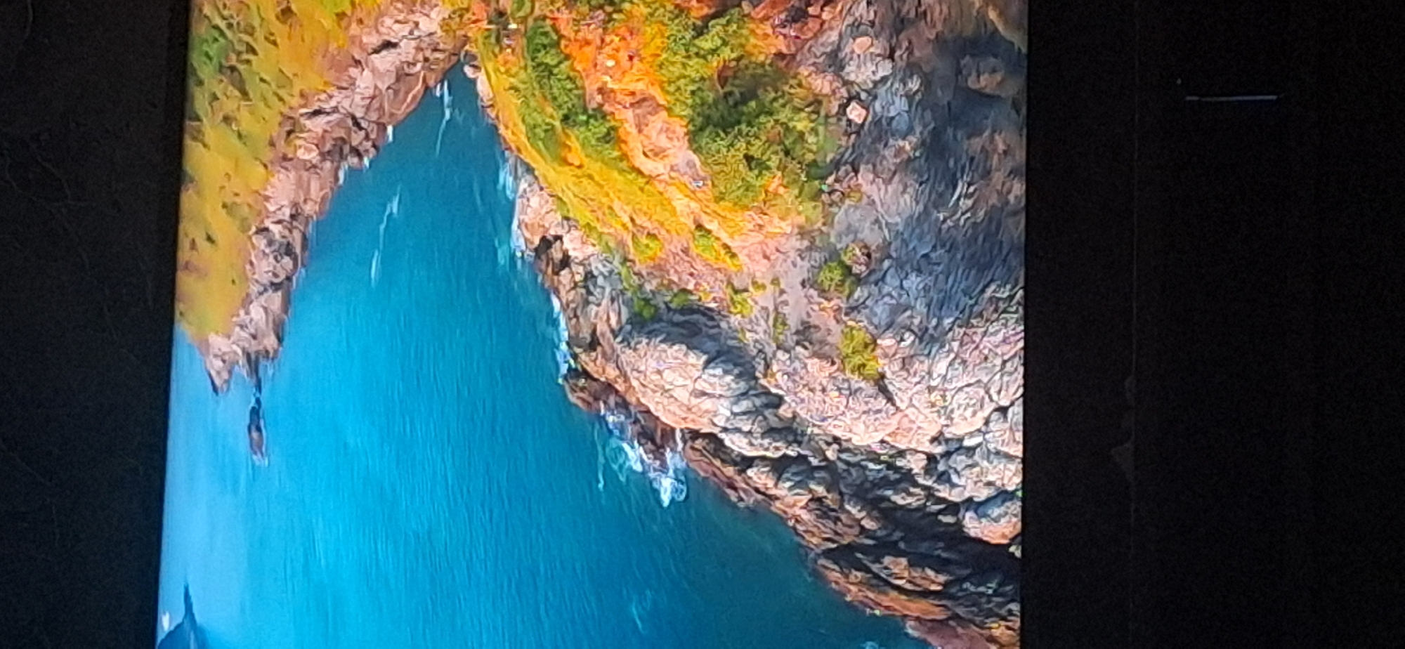 The image shows a rocky coastline with blue water lapping against it. The land is covered in green and yellow vegetation. There are small figures visible on the grassy slopes, suggesting people are present. The image appears to be a still from a video or a photograph of a natural landscape. There is no information or visual evidence to suggest this location is in the city of Bila Tserkva, Ukraine, or any other specific city. The scene depicts a coastal environment.