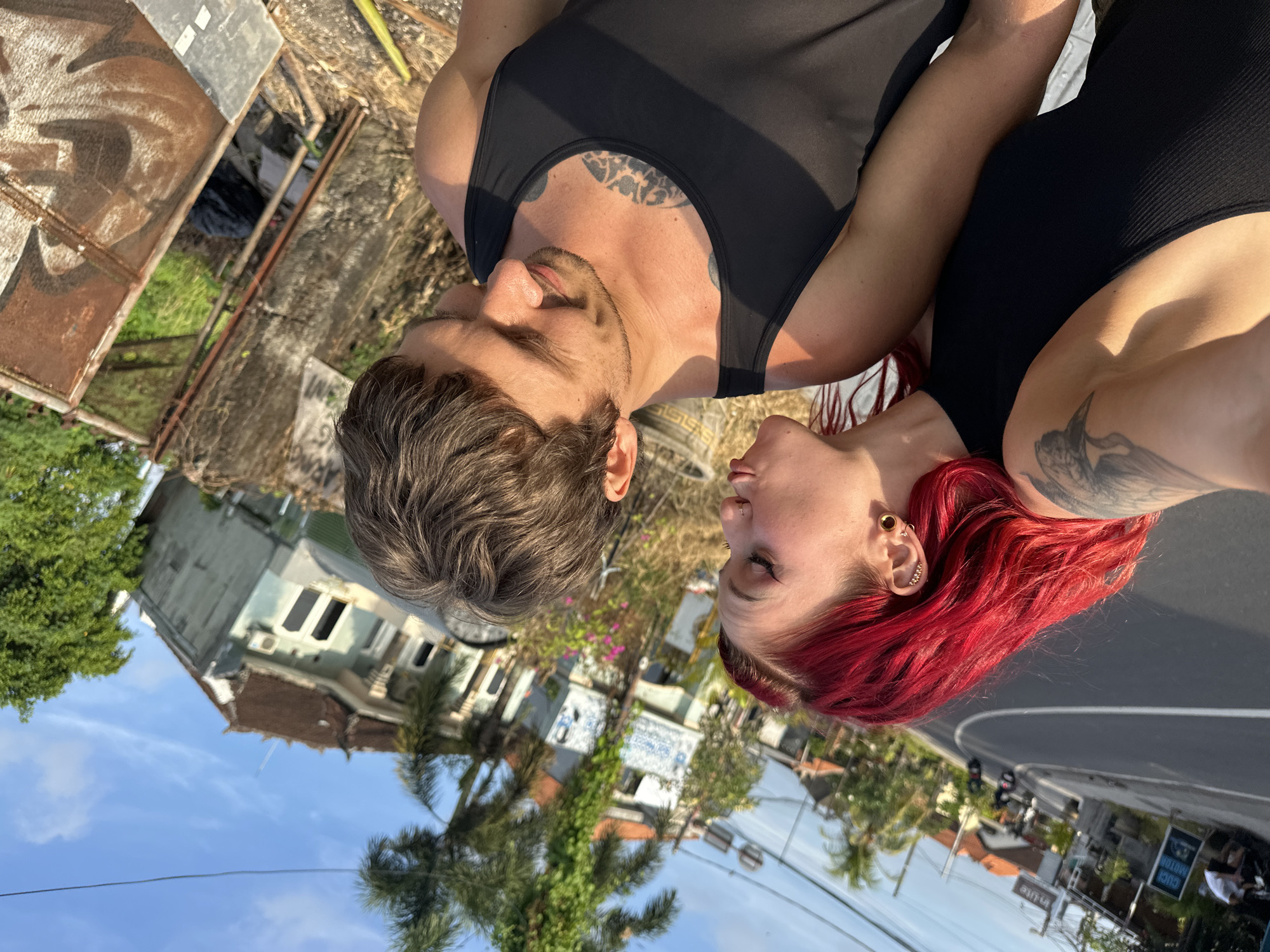The image, oriented 90 degrees clockwise, depicts a close-up portrait of two individuals, a man and a woman, facing each other with their heads tilted downwards. The man, on the left, has short to medium brown hair and a short beard. He wears a black tank top, revealing a tattoo on his upper chest. The woman, on the right, has vibrant red hair and a fair complexion. She has a nose ring, multiple ear piercings including a large gauge, and a tattoo on her right arm/shoulder. She is wearing a black ribbed sleeveless top. Both individuals have their eyes closed or looking down, suggesting a moment of intimacy or reflection.
The background reveals an outdoor setting in North Kuta, Indonesia. To the left of the man, there are some worn structures, possibly corrugated metal fencing, and green foliage. Between the two, some small pink and purple flowers are visible. To the right of the woman, a paved road or path is present, curving into the distance. Beyond the road, there are residential buildings, palm trees, and a clear blue sky with sparse clouds. The lighting is soft and warm, characteristic of late afternoon or early morning. A clear location cue is visible text on a sign by the road in the background, which reads "CUCI MOTOR" and "MATIC", indicating a motorcycle wash in Indonesian.