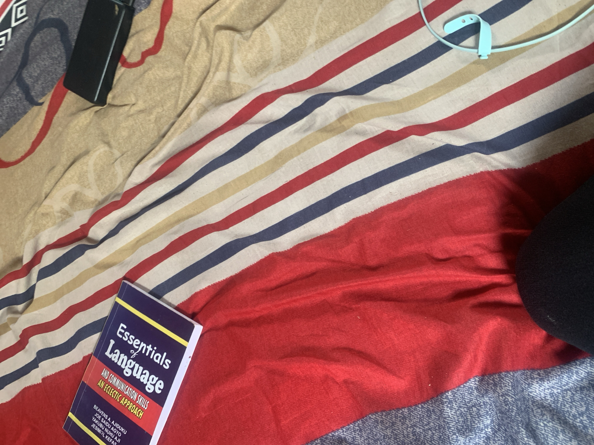 A book titled "Essentials of Language" with the subtitle "An Eclectic Approach and Communication Skills" is placed on a striped bedspread. The bedspread features red, white, blue, and yellow stripes. To the right of the book, a black power bank is visible, and further to the right, a light blue earbud loop rests on the bedspread. In the lower right corner, a person's black leg is visible. The image appears to be taken indoors, possibly in a bedroom, and the setting does not provide any specific clues about its location in Jauro Yinu, Nigeria.