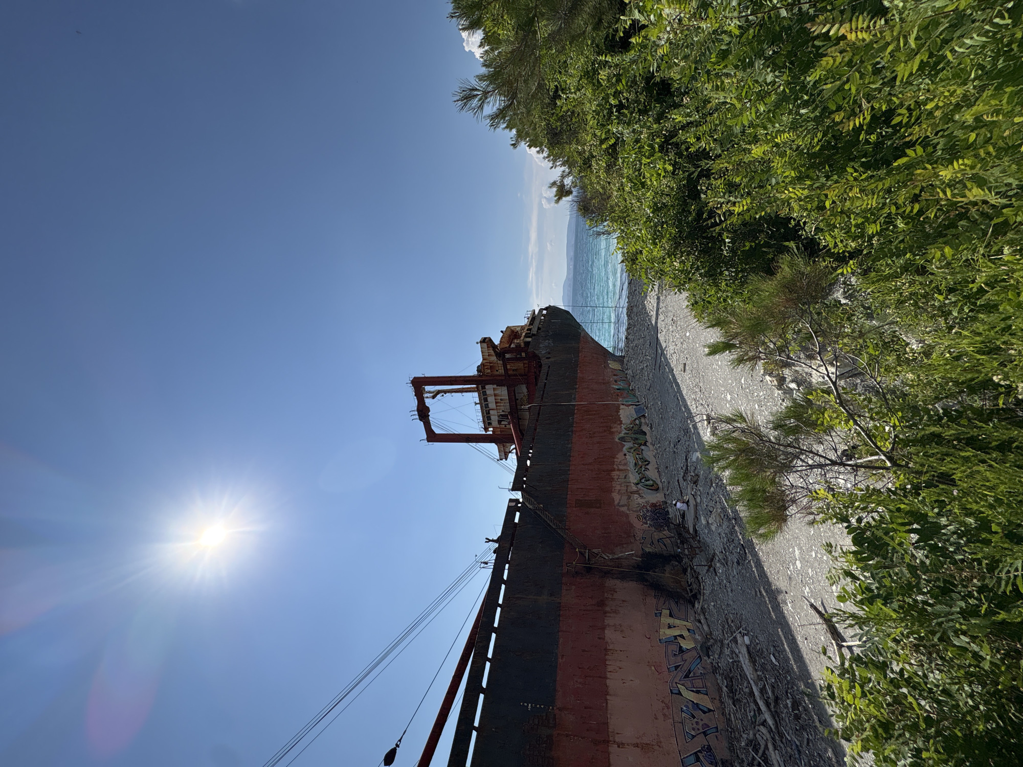 A large, rusted cargo ship is beached on a pebble shore, its hull painted in sections of red and dark gray, adorned with graffiti. The ship occupies the majority of the frame, leaning towards the right. The sea, a vibrant turquoise, is visible behind the ship. Lush green foliage frames the scene on the right side, partially obscuring a distant shoreline and hills. The sky above is a clear, bright blue with the sun prominently featured in the upper left quadrant, casting a bright glare. The environment suggests a sunny day in an outdoor location, likely coastal. The image appears to be taken from a slightly elevated position, looking down towards the ship and the water.