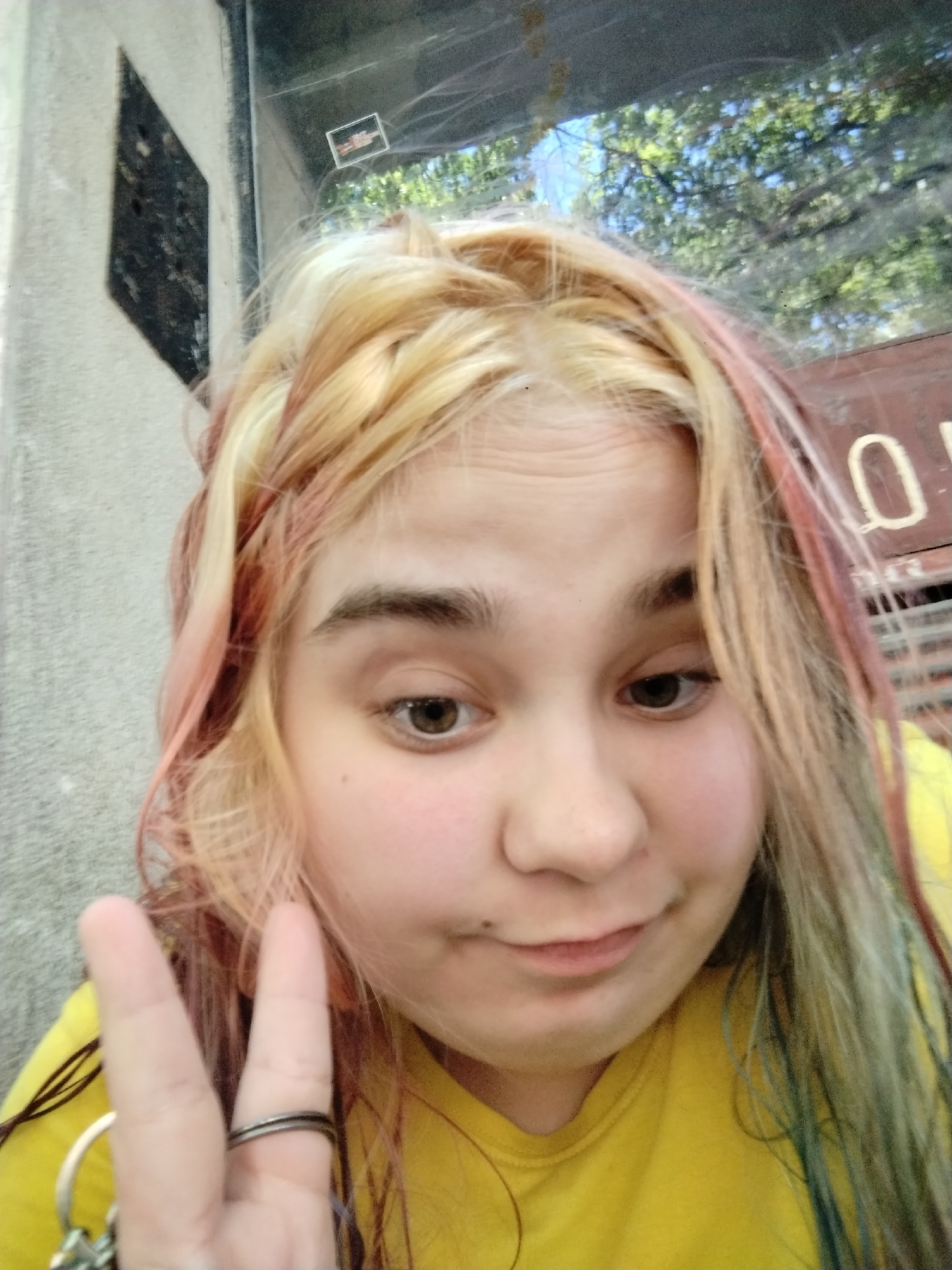 A close-up selfie of a person with light-colored hair, some strands dyed pink, green, and blonde. The person is wearing a yellow t-shirt and making a peace sign with their left hand, which also holds keys. The person has brown eyes, fair skin, and a mole near their mouth. The background is a mix of outdoor and indoor elements. A light gray wall is partially visible on the left, with a dark, diamond-shaped design. Behind the person, there appears to be a window reflecting trees and foliage, suggesting an outdoor setting. The reflections indicate that the person might be inside a bus or a building with windows. The location in Plovdiv, Bulgaria, is unconfirmed based on the image alone.