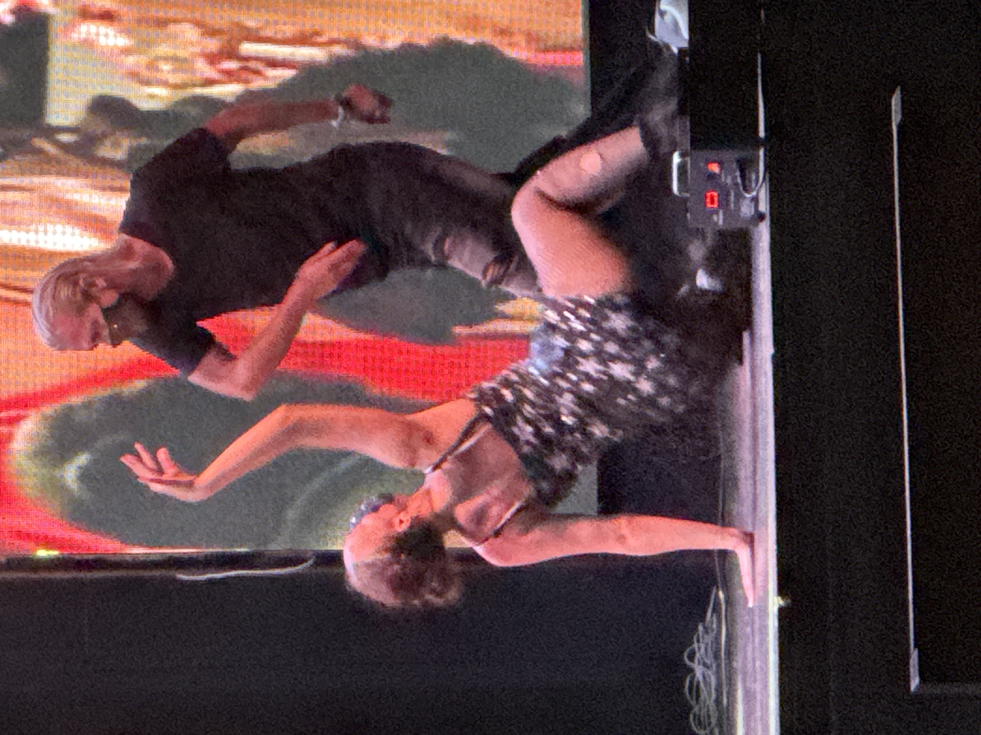 The image shows a man and a woman dancing on a stage with a brightly lit background. The woman is in the foreground, wearing a sparkly black dress and is in a low, dynamic pose on the stage, with her body angled and one leg bent. Her arms are raised and her hand is open. The man is behind her, wearing a black t-shirt and dark pants, and appears to be leaning down, interacting with her. He has blonde hair and is wearing a mask. The stage floor is visible, with some electrical equipment to the right of the woman. The overall scene suggests a performance or event taking place in an indoor venue. There is no information in the image to suggest it is located in Manavgat, Turkey.