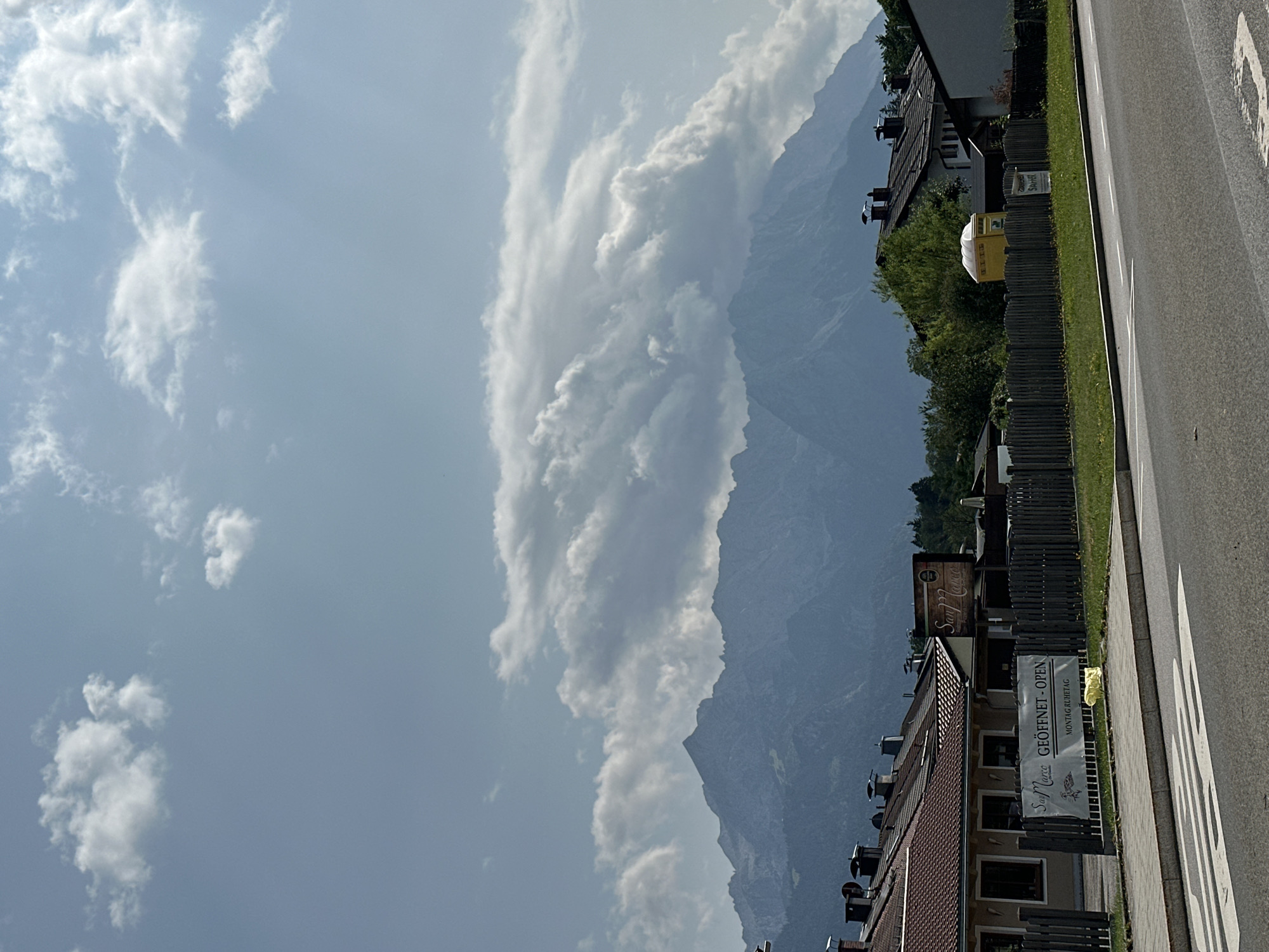 The image shows a street view in Garmisch-Partenkirchen, Germany, with residential buildings and a restaurant in the foreground, and the Zugspitze mountain range in the background. The sky is partly cloudy with sunlight breaking through. In the foreground, there is a street with a pedestrian crossing. To the right of the street, there is a wooden fence and a grassy area. Behind the fence are several residential buildings with red-tiled roofs and chimneys. A banner on one of the buildings reads "San Marco GEÖFFNET - OPEN MONTAG RUHETAG". Further back, there are trees and the imposing mountain range. A yellow portable toilet is visible behind the fence. The scene appears to be calm and depicts a typical day in a Bavarian town.