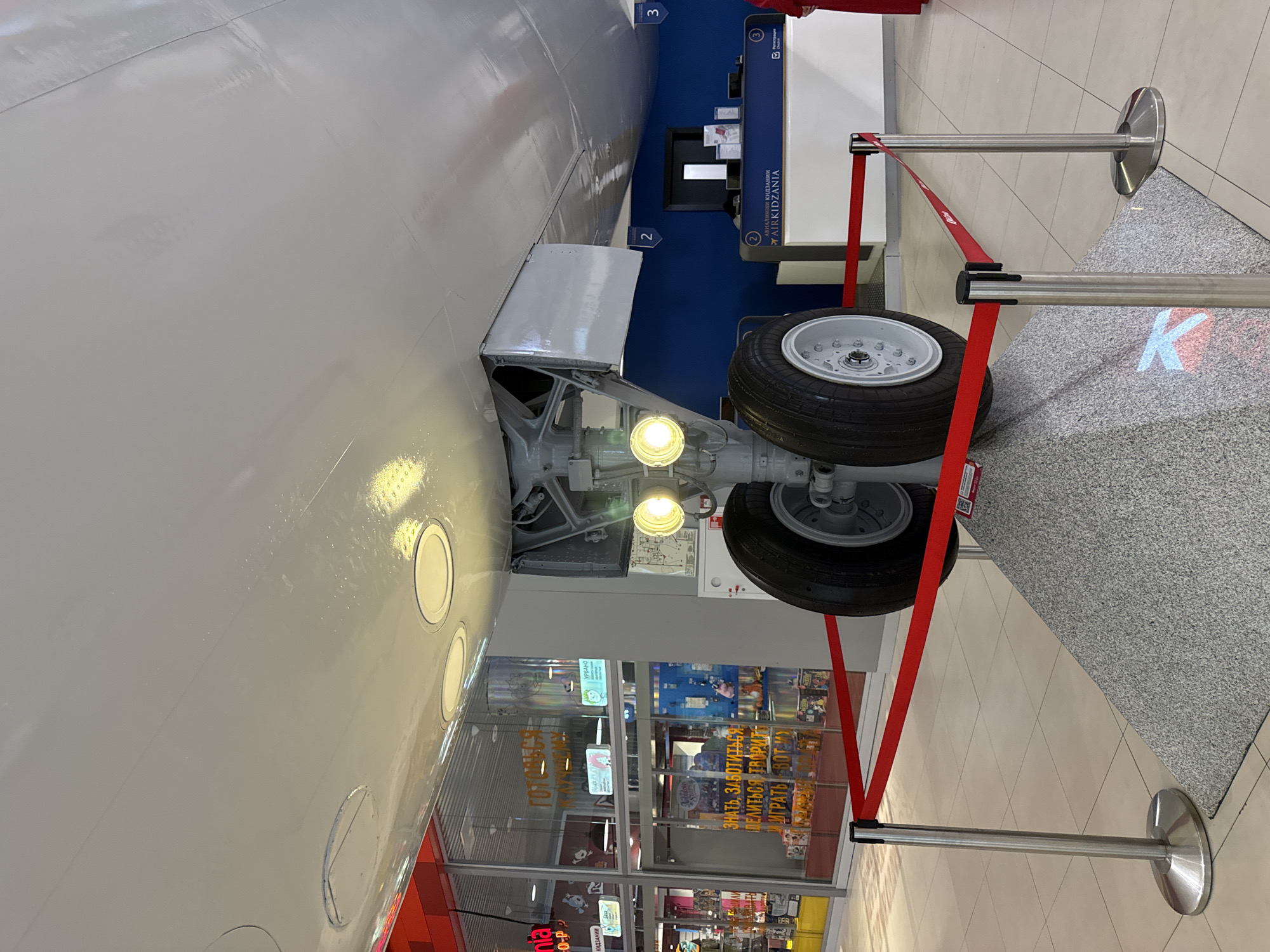 This is a view of the landing gear of an aircraft, likely as part of an exhibit. The landing gear is in the foreground, with two wheels visible. It is behind a red rope barrier. Behind the landing gear, a shop can be seen with colorful displays of merchandise. The text in Russian is visible on the shop windows. The landing gear appears to be installed inside a building or museum, judging by the smooth white walls and the presence of signs indicating "AirKidzania" and numbered panels (2 and 3), suggesting it's part of an educational or entertainment exhibit. A tiled gray floor can be seen to the right of the red barrier. The exhibit is located in Khimki, Russia.