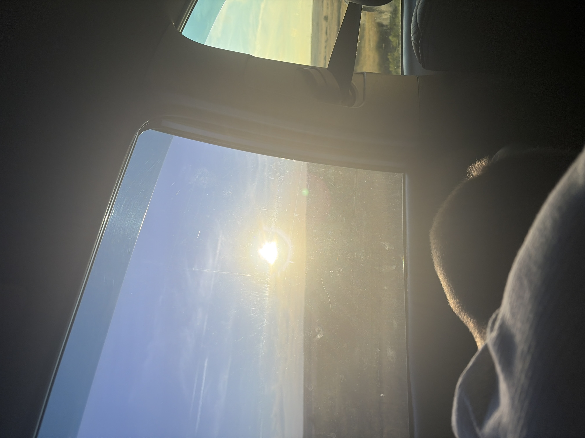The image depicts a view from inside a vehicle, looking out the passenger side window towards a sunlit landscape. The sun is positioned in the upper center of the frame, emitting a bright glare that washes out details in the sky and on the window. The sky is a gradient of blue and yellow, with some faint clouds or contrails visible. The landscape outside appears to be a flat, grassy field or prairie with some scattered vegetation and a distant treeline. The window itself is smudged and scratched, with reflections from the interior of the car visible. In the foreground, a portion of the car's interior is visible, including a seat and a person's head or shoulder covered by a light-colored fabric. The overall lighting suggests it is either sunrise or sunset. The scene is consistent with a rural or exurban environment, and the details suggest the image was taken within the city of Belogorki, Russia.
