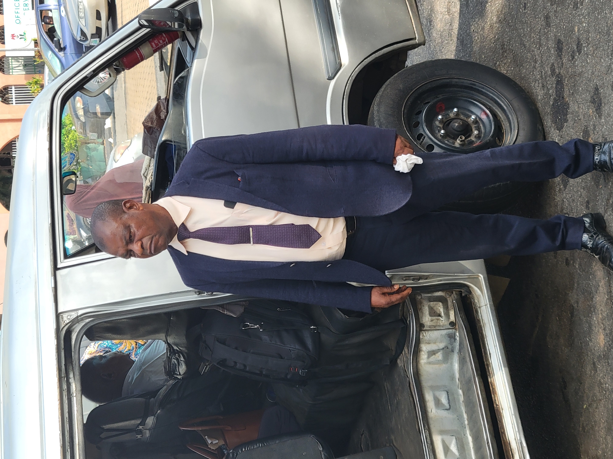 A middle-aged African man in a dark suit, light shirt, and patterned tie stands beside a silver van. He is holding a white tissue in one hand and touching the van's open door with the other. Inside the van are bags and another person seated in the back. The van is parked on a paved street. In the background, other vehicles are visible, along with a building with the partial text "OFFICE OF ENVIRONMENTAL SERVICES". The location is likely in the city of Abuja, Nigeria.