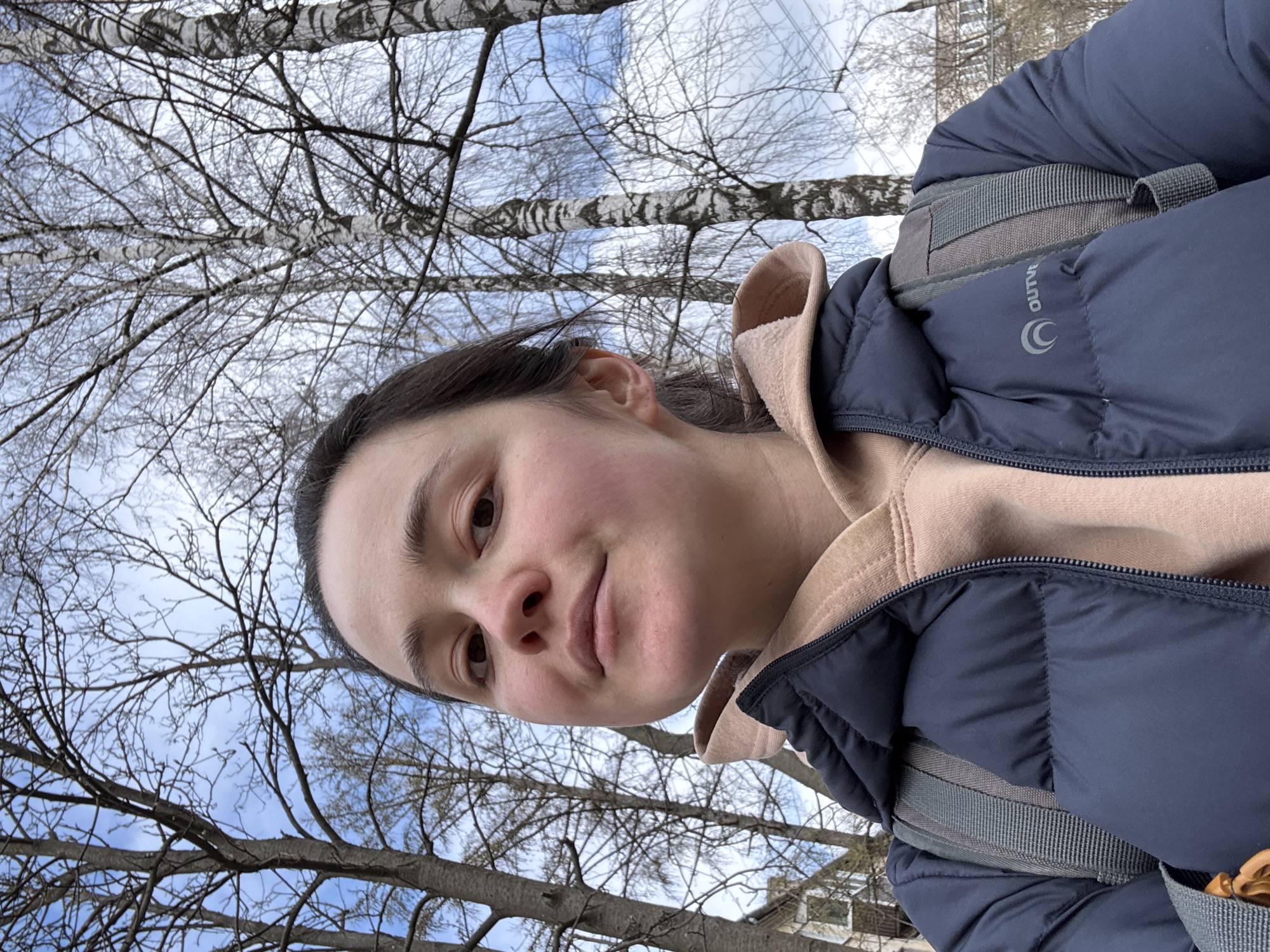 This is a close-up, upward-facing portrait of a young adult female, taken outdoors in Izhevsk, Russia. The subject has dark hair pulled back, fair skin, and is looking towards the camera with a subtle, closed-mouth smile.
She is dressed in a dark blue puffer jacket worn over a light beige or brown hooded sweatshirt. A grey backpack strap is visible over her right shoulder (viewer's left). On the backpack strap, the partial text "OUTV" along with a crescent moon logo is clearly visible.
The background consists of a dense canopy of bare tree branches, suggesting late winter or early spring. Several tree trunks with characteristic white and black bark, likely birch trees, are prominent across the upper frame. The sky behind the branches is bright blue with scattered clouds. In the far distance, through the network of branches, faint structures that appear to be buildings can be seen. The lighting indicates it is daytime, possibly late morning or early afternoon. The overall impression is one of calm, capturing a moment outdoors amidst nature.