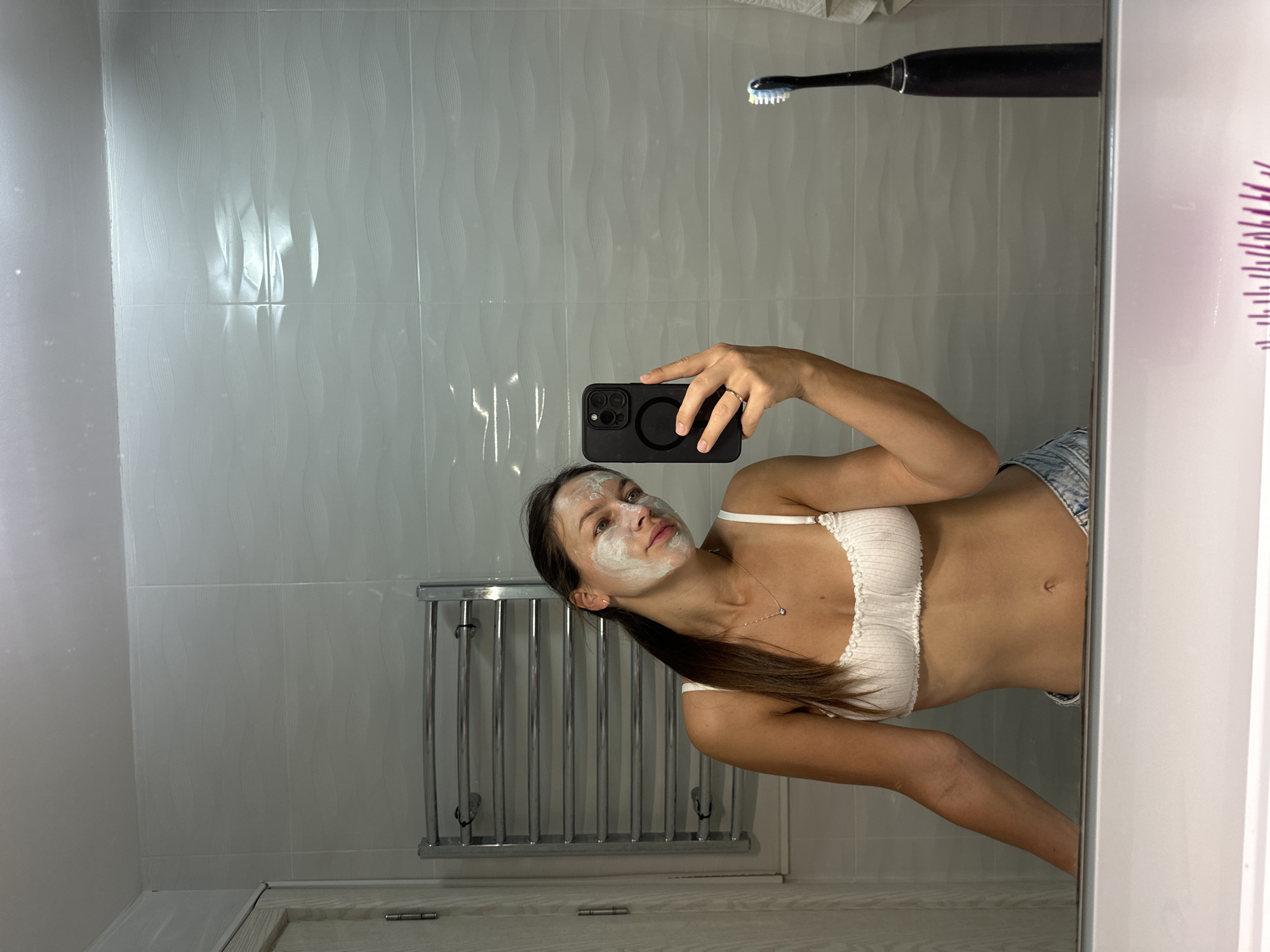 A young woman with a white facial mask applied to her face takes a mirror selfie in a bathroom. She is wearing a white bralette and denim shorts. To her left, a chrome towel rack is visible. To her right, a black electric toothbrush stands upright. The bathroom walls are tiled with a wavy pattern. The reflection shows the upper part of a dresser or vanity in the foreground. The location is identified as a bathroom in Bečići, Montenegro.