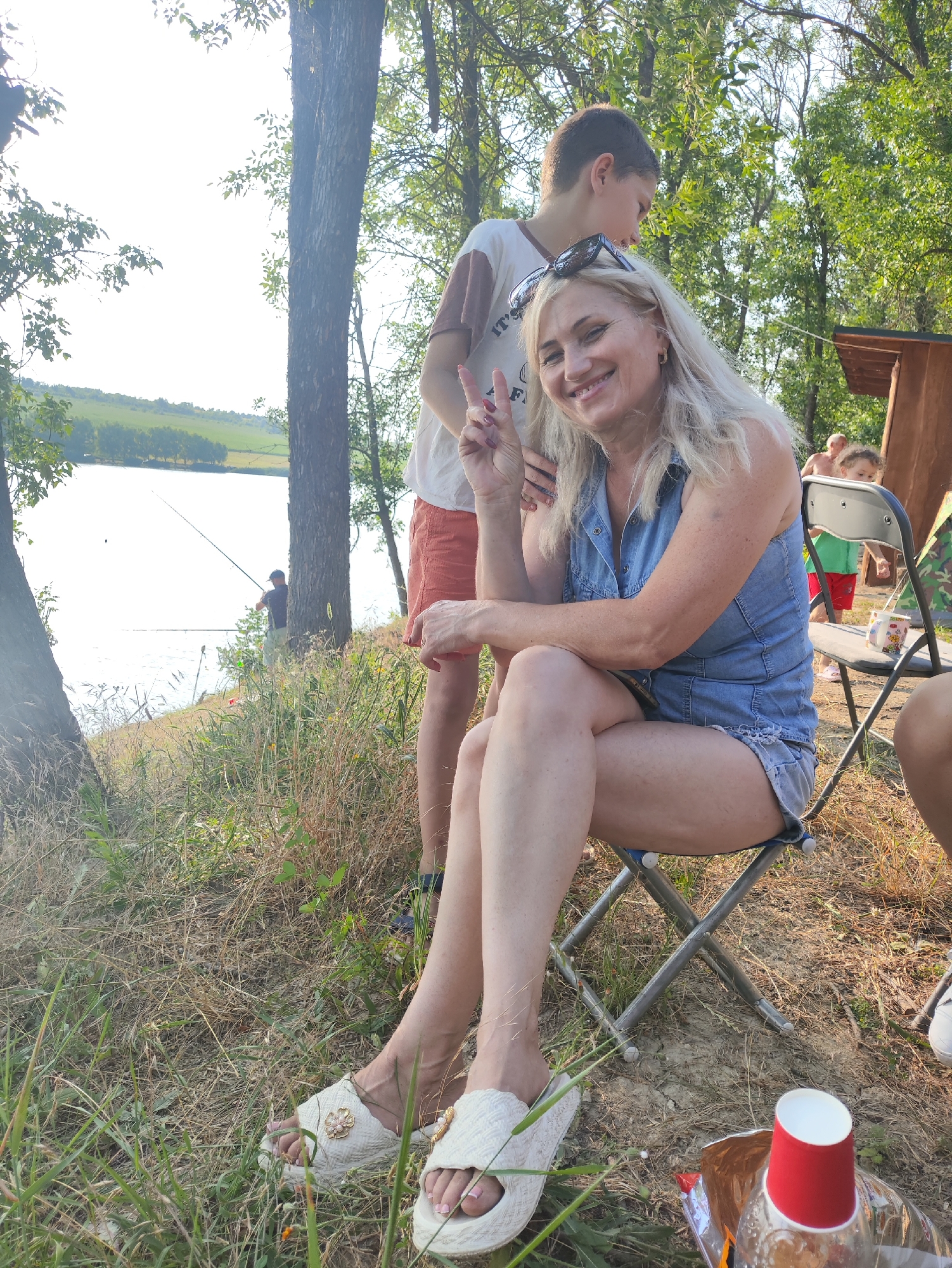 In Găleștii Noi, Moldova, a woman with blonde hair and sunglasses on her head is sitting on a folding chair and smiling at the camera, holding up two fingers in a peace sign. She is wearing a denim shirt and shorts. Behind her, a young person in an orange-and-white t-shirt and orange shorts stands. In the background, a lake is visible, with a person fishing by the water. Other individuals and a wooden structure are also present in the scene. The setting is outdoors, with trees and dry grass indicating a natural environment. A red paper cup and a bottle are on the ground in the foreground.