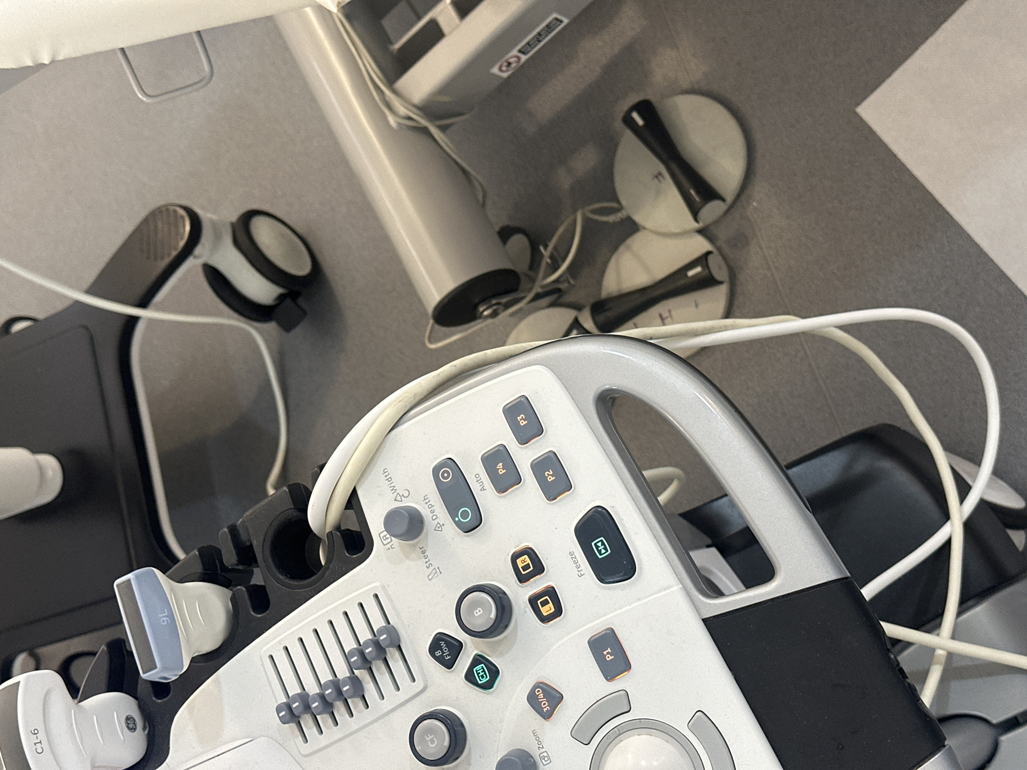 A high-angle shot shows the top portion of a GE ultrasound machine in a medical setting. The machine's control panel, silver and white with numerous buttons, knobs, and a handle, dominates the lower half of the frame. Various probes are attached to the machine, including labeled "C1-6" and "9L". Wires and cables are strewn around the machine. In the background, cylindrical objects and round electrodes can be seen on a gray surface, possibly a countertop. The environment appears sterile, suggesting a hospital or clinic setting. There's no indication of the city of London, United Kingdom.
