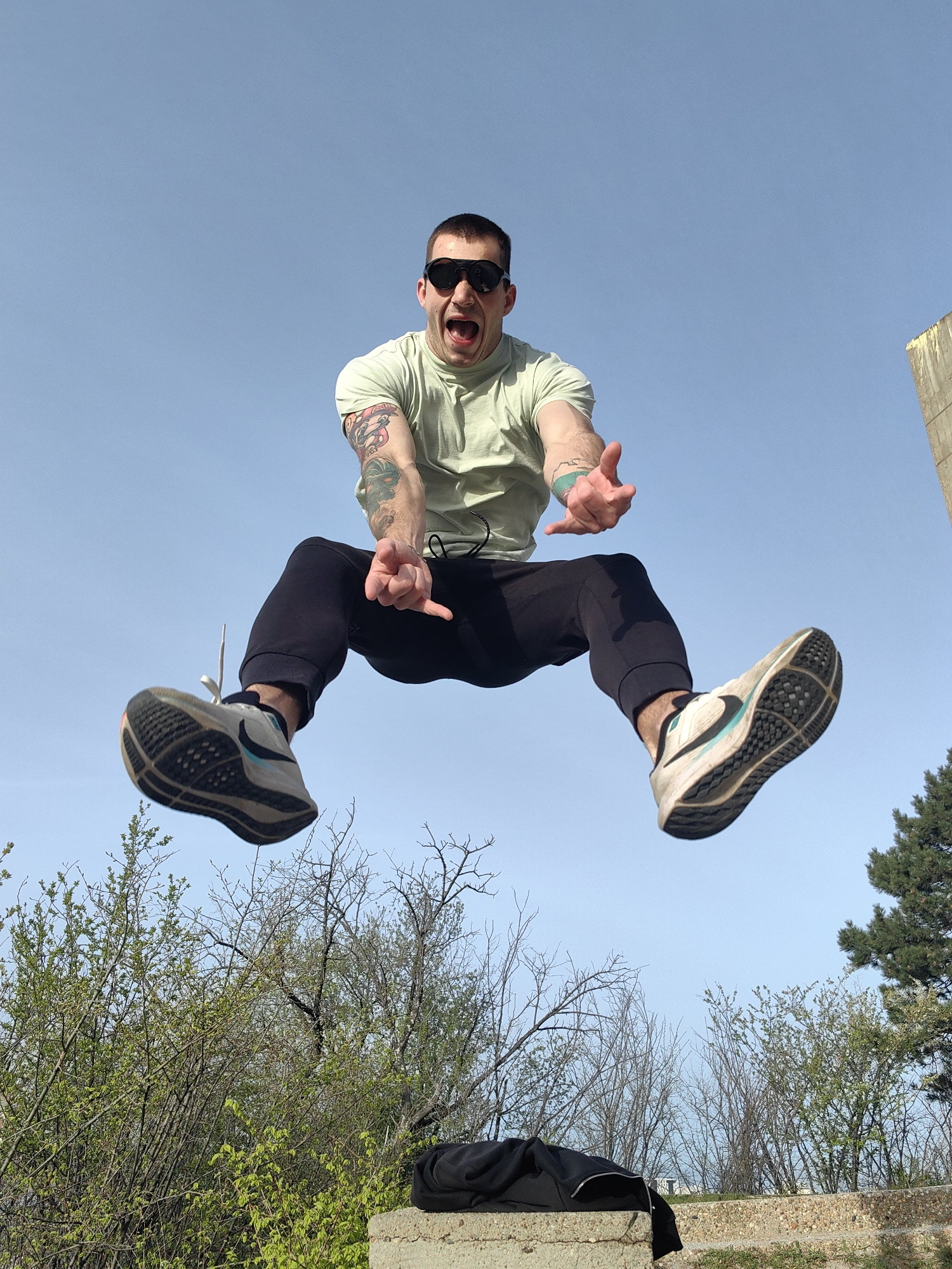 A man wearing a light green t-shirt, black pants, black sunglasses, and athletic shoes is captured mid-air against a clear blue sky. He is jumping with his legs bent and his arms outstretched. He has tattoos on his arms and is shouting or yelling with his mouth open. Below him, there is green foliage and bare trees. A concrete structure is present on which a black garment is placed. To the right, there is a tall conifer tree and a concrete corner of what appears to be an unfinished building. The location is likely a park in Varna, Bulgaria, judging by the vegetation and the architectural elements present in the background.