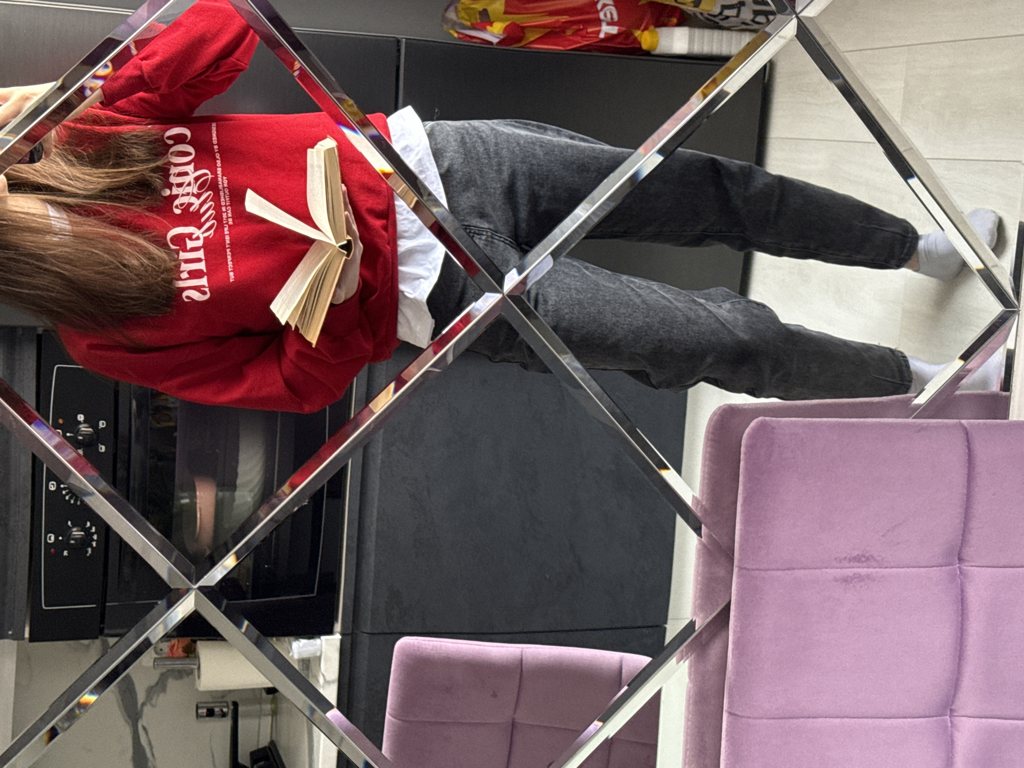 A person is seen through a wall mirror composed of diamond-shaped sections. The person, wearing a red "Coffee Clip" sweatshirt and gray jeans, sits with legs stretched out. They are holding a book open in their hands. In the background, there's a black cabinet, what appears to be a kitchen oven, and lavender chairs. There are no identifiable features that indicate the city, but the setting appears to be indoors, possibly in a kitchen area.