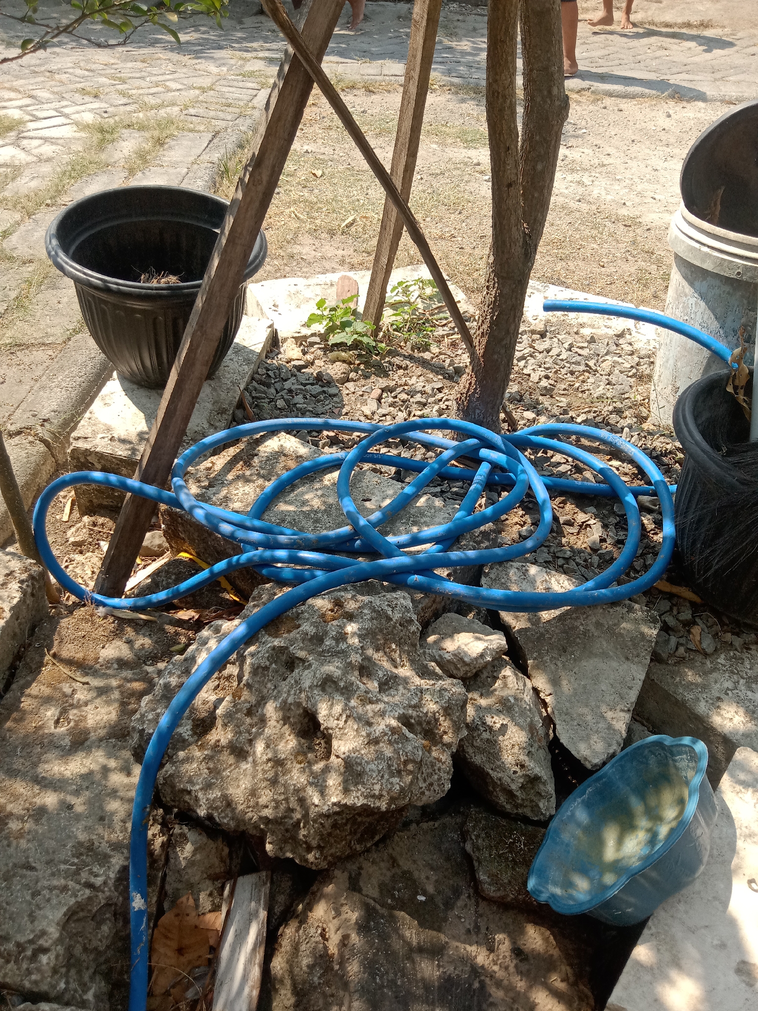 In Stiaji, Indonesia, a blue garden hose is coiled on a rocky surface next to a tree and several large rocks. A black plastic planter is to the left, and a grey plastic bin is to the right. The ground is covered in small stones and dirt. Sunlight casts shadows on the scene. In the background, two people are visible standing on a paved pathway.