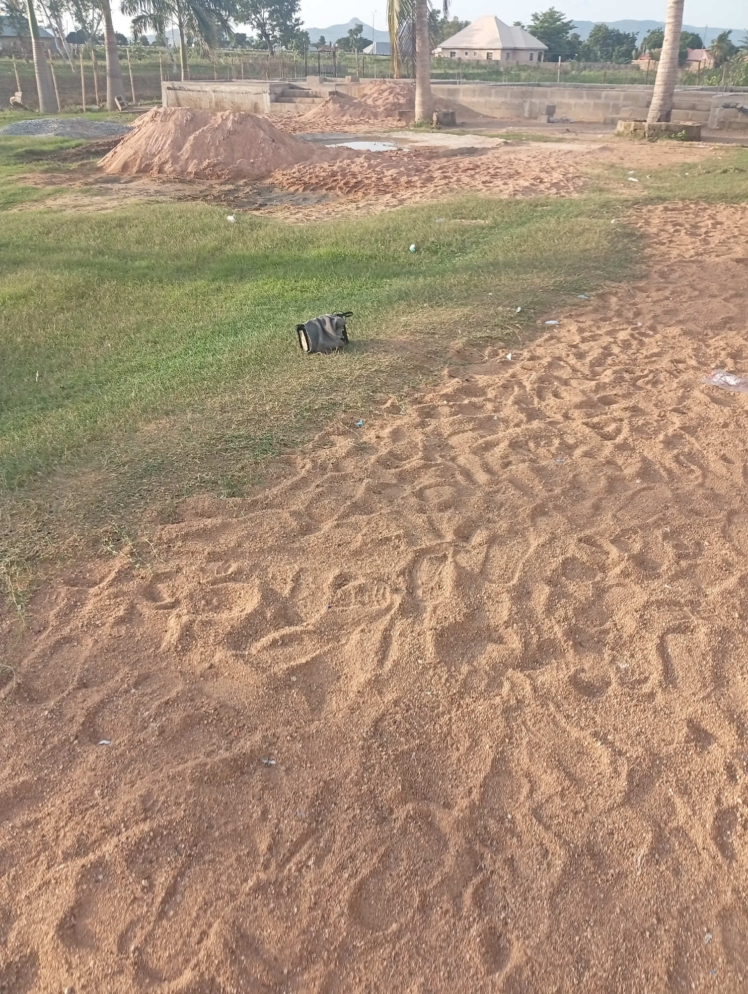 This media file depicts an outdoor scene in Jalingo, Nigeria. The foreground is dominated by sandy ground with visible footprints and tire tracks, suggesting recent activity. In the mid-ground, a patch of green grass transitions into the sandy area. A dark-colored bag, possibly a duffel bag, rests on the grass.
In the background, the scene appears to be a construction site. Piles of sand and gravel are visible, along with the beginnings of a concrete structure, possibly a building foundation. Several palm trees are scattered throughout the area, some appearing mature and tall, while others are younger. A building with a light-colored, pitched roof is situated further in the background, with more residential or commercial structures visible beyond that. The sky is partly cloudy, and the lighting suggests it is daytime, likely late afternoon or early morning due to the soft light. The overall impression is one of an ongoing construction project in a semi-rural or developing area. No people are clearly visible in the frame.