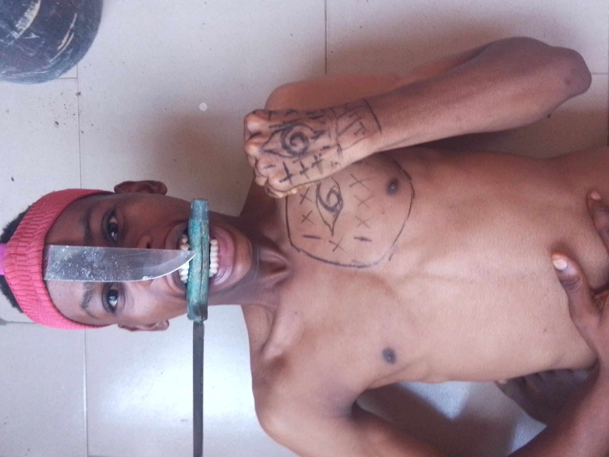 A young person, appearing to be male and bare-chested, is depicted from the waist up. The image is rotated 90 degrees clockwise. The person is holding a large, sharp metal knife blade in their mouth, clenched between their teeth. The handle of the knife, which appears to be green or blue-green, is attached to a long, thin, dark metal rod that extends downwards out of the frame. The person wears a red knitted headband on their head and looks directly at the camera with wide eyes and an open mouth, revealing their teeth.
Black, eye-like symbols with surrounding lines and crosses are drawn onto their right clenched fist and on their chest. The background shows a light-colored tiled surface, likely a floor or wall, with a dark, round object partially visible in the upper left corner. The scene appears to be indoors.