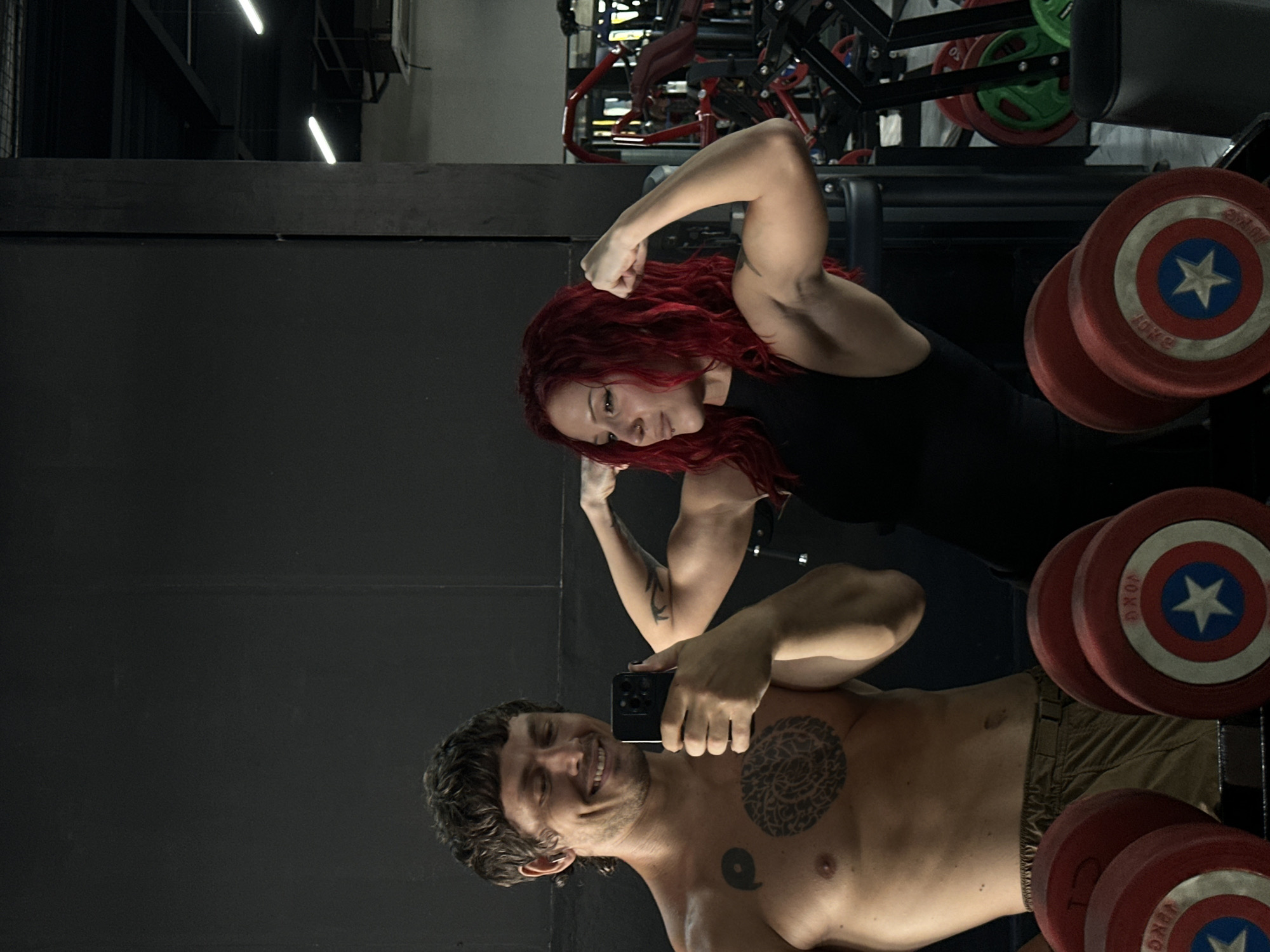 A man and a woman are taking a selfie in a gym in North Kuta, Indonesia. The man is shirtless with a tattoo on his chest and is holding a phone to take the picture. He is smiling. The woman is wearing a black tank top and has red hair. She is flexing her arm. There are weight plates with a Captain America design in the foreground and gym equipment in the background.