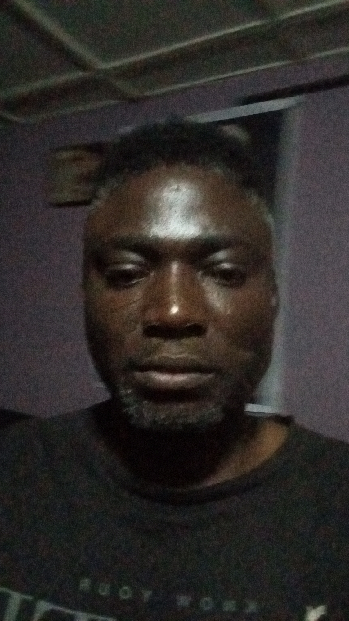 A close-up portrait of a dark-skinned man with short, dark hair and a grey beard, likely indoors. His skin appears shiny and slightly sweaty. He is wearing a dark-colored t-shirt. The background is blurry, showing a textured, dark purple wall. The location and specific context related to Sabon Tasha, Nigeria, are not identifiable from the image.