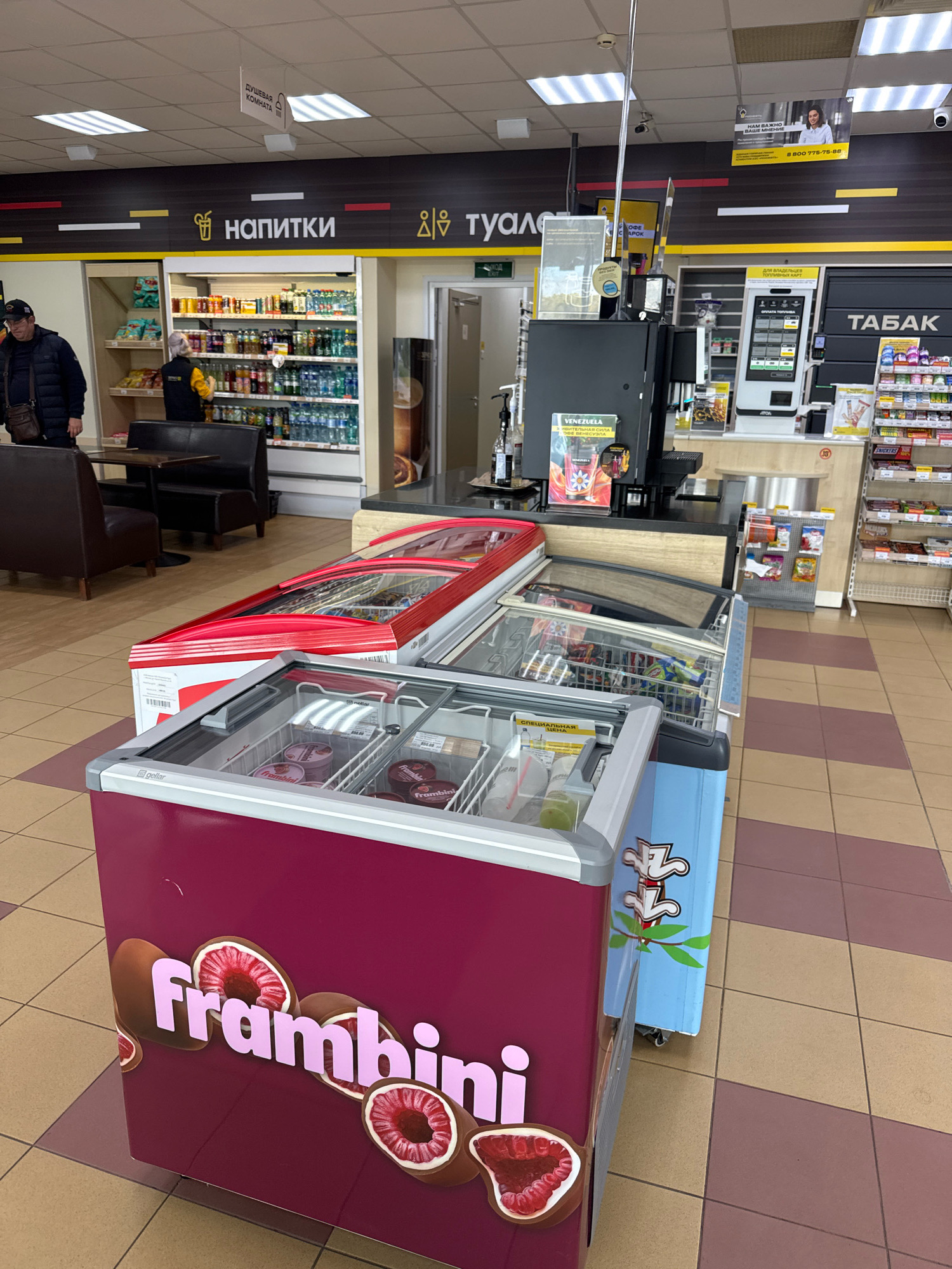 The image shows the interior of a convenience store, likely a gas station convenience store. In the foreground, there are two glass-top freezers filled with ice cream or frozen desserts. The freezer on the left has a maroon-colored side with the text "frambini" and images of raspberries. The freezer on the right is light blue. Behind the freezers, there is a section of shelving with various products. Further in the background, there are refrigerators stocked with drinks, a coffee machine, and a payment terminal. A sign above a section of shelving reads "НАПИТКИ" (DRINKS) and "ТУАЛЕТ" (TOILET). A person in a black jacket is standing near a table and chairs in the seating area. Another person in a yellow vest is stocking shelves. The store has a checkered tile floor and a drop ceiling with fluorescent lighting. There are also advertisements on the walls. The image appears to be taken from a standing perspective. The location of Novoselitsy, Russia is not visually evident from the image.