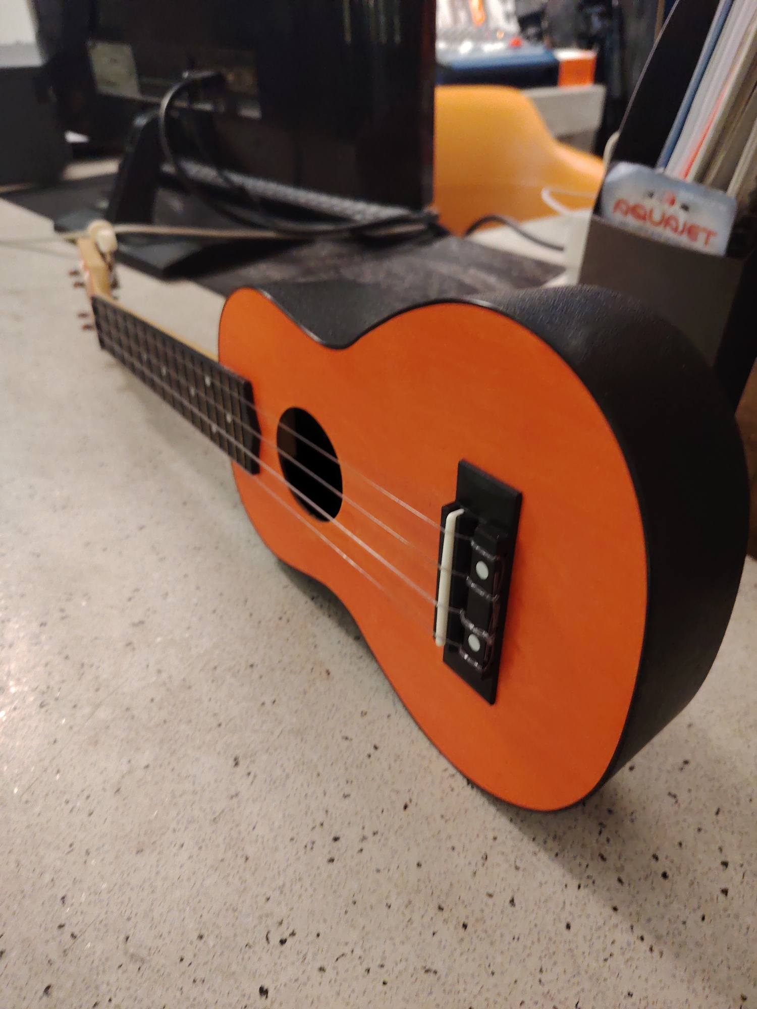 An orange and black ukulele is positioned on a light-colored, speckled floor within an indoor environment in Unknown, Unknown. The ukulele's soundboard is orange, contrasting with its black sides, back, and fretboard. Four white strings are strung from the partially visible headstock, over a black bridge equipped with a white saddle, and across a circular soundhole on the instrument's face. The floor exhibits a light grey or off-white base with scattered dark speckles. The background is blurred, revealing the rear of a dark electronic display unit with connected cables, a curved orange object, and a dark desk organizer. Within the organizer, a white item bears the red inscription "AQUAJET". No individuals are present in the scene.