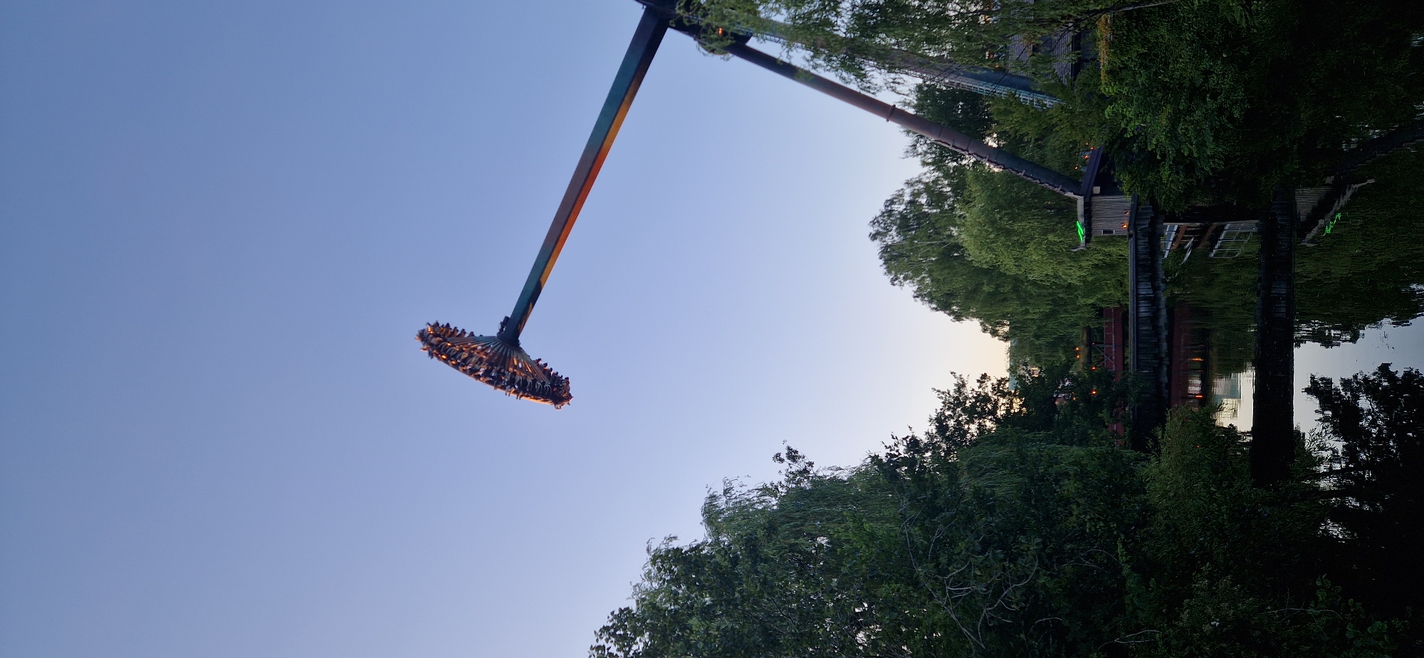 The image shows a large pendulum ride at an amusement park, possibly Liseberg in Gothenburg, Sweden. The ride, with passengers seated on a circular platform at the end of a long arm, is tilted upwards against a clear blue sky. Green trees frame the scene on the left and right, and a body of water with reflections of trees and structures is visible in the foreground. A bridge crosses the water in the mid-ground. The lighting suggests it is late afternoon or early evening.