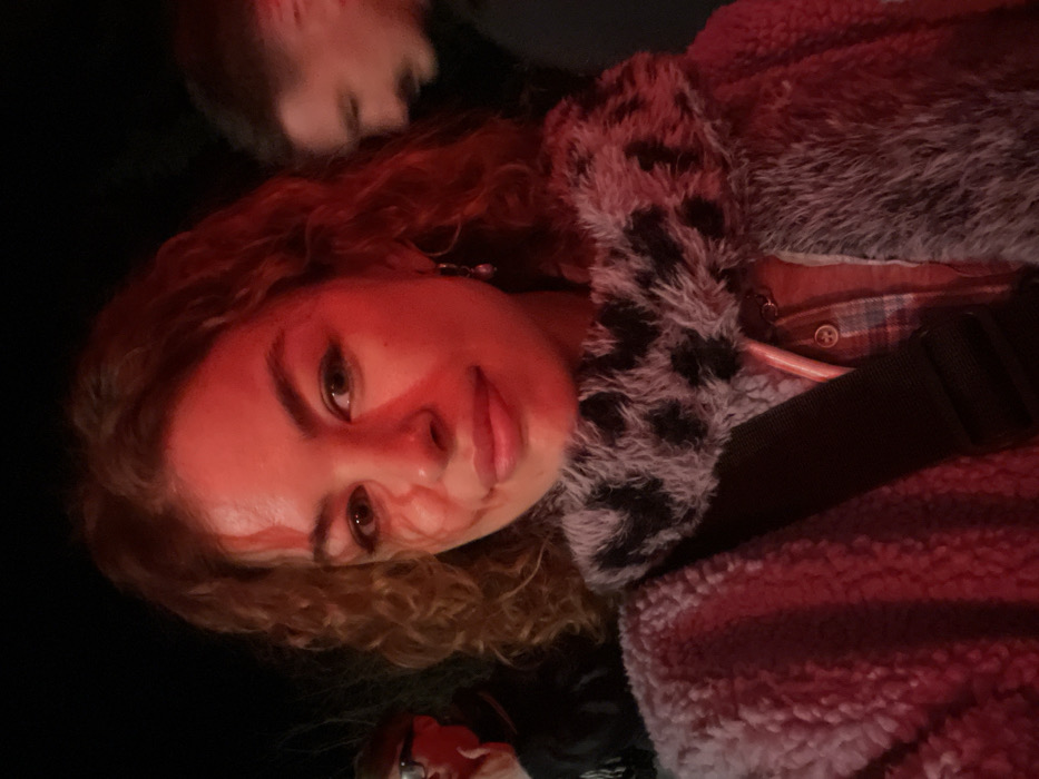 A young woman with curly red hair looks at the camera under red lighting. She wears a fuzzy leopard print collar and a plaid shirt. A black strap crosses her body. A person with short dark hair and glasses is partially visible in the bottom corner, while another individual is barely seen at the top. The background is mostly dark.