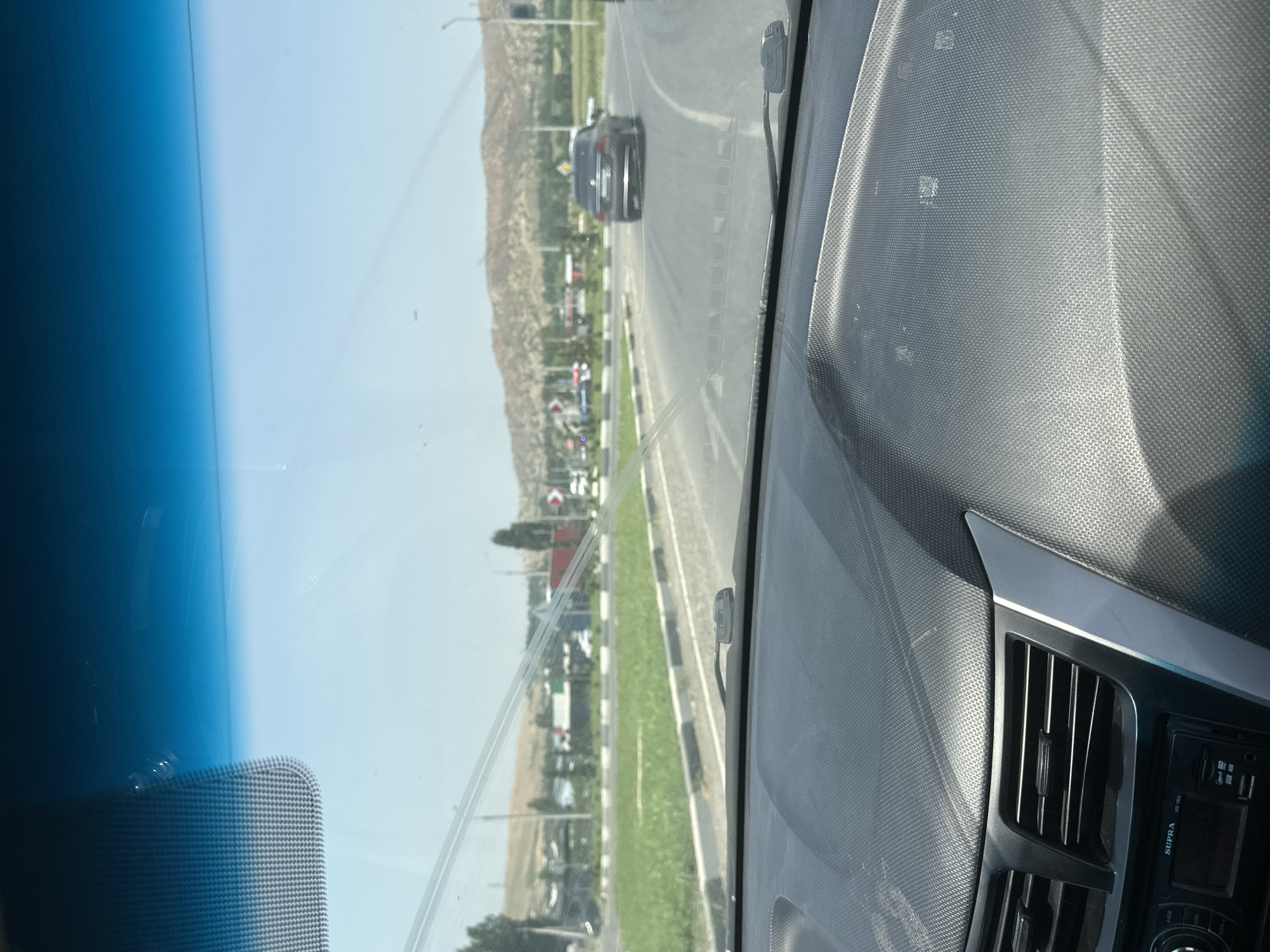 The image is taken from inside a vehicle, looking out through the windshield. The view shows a road with a car driving ahead, likely on a highway or main road. There are grassy areas and trees on the side of the road, with hills or mountains in the background under a bright, clear sky. The dashboard of the car is visible in the foreground, with an audio system and air vents. There are some water streaks or smudges on the windshield. The scene appears to be in Suzak, Kyrgyzstan.