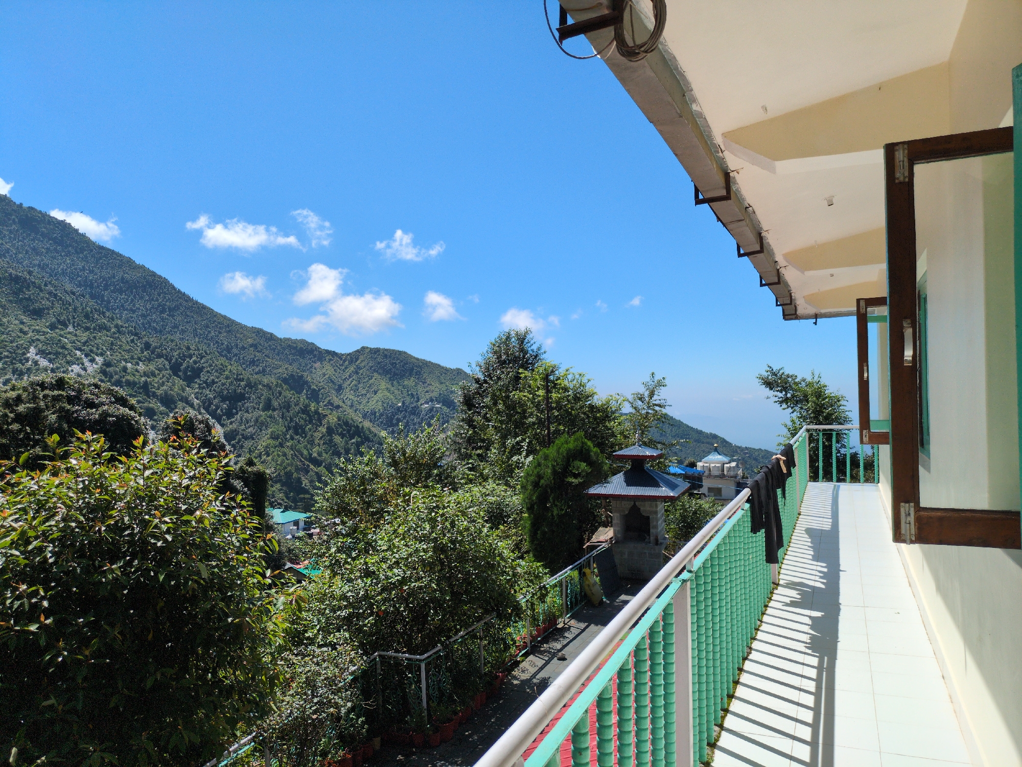 A sunny day with a clear blue sky and scattered clouds above a lush green mountain range. The viewpoint is from a balcony with a white tiled floor and a green railing. A person is standing near the railing on the balcony, facing away from the camera and looking out at the view. Some black clothing is hanging on the railing, casting long shadows across the balcony floor. In the distance, there are trees and buildings, suggesting a settlement nestled within the mountains. The overall scene depicts a serene and scenic mountain landscape. The location is in the city of Haini, India.