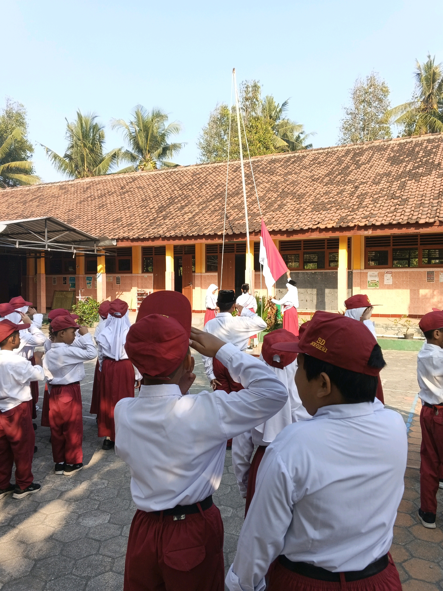 A group of elementary school children in uniform are participating in a flag-raising ceremony in front of a building with a terracotta tiled roof. The children are wearing white shirts and maroon pants or skirts, and maroon caps. Some of the children are saluting, while others are looking towards the flag. A flag with red and white stripes is being raised on a flagpole. The scene appears to be set in a school courtyard with palm trees visible in the background. The location is in Glagah, Indonesia.