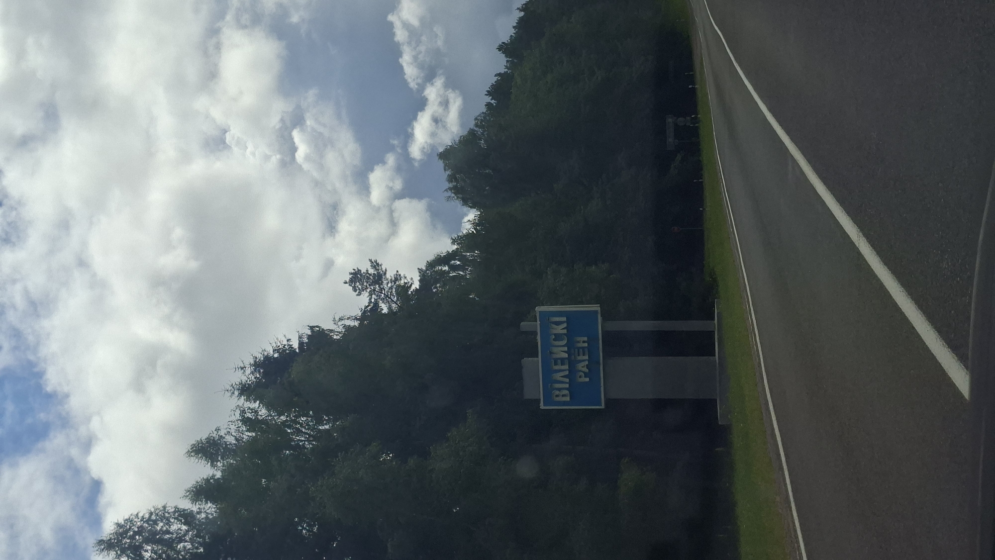 The image shows a road in a rural area of Belarus. On the left side of the road, there is a blue sign with white text that reads "ВІЛЕЙСКІ РАЁН". The sign is mounted on a gray pillar. Behind the sign, there is a dense forest. The sky is partly cloudy with white and gray clouds. The road is asphalt and has white lane markings. There are no people visible in the image, but a car can be seen in the distance on the right side of the road. The overall impression is one of a quiet, natural landscape.