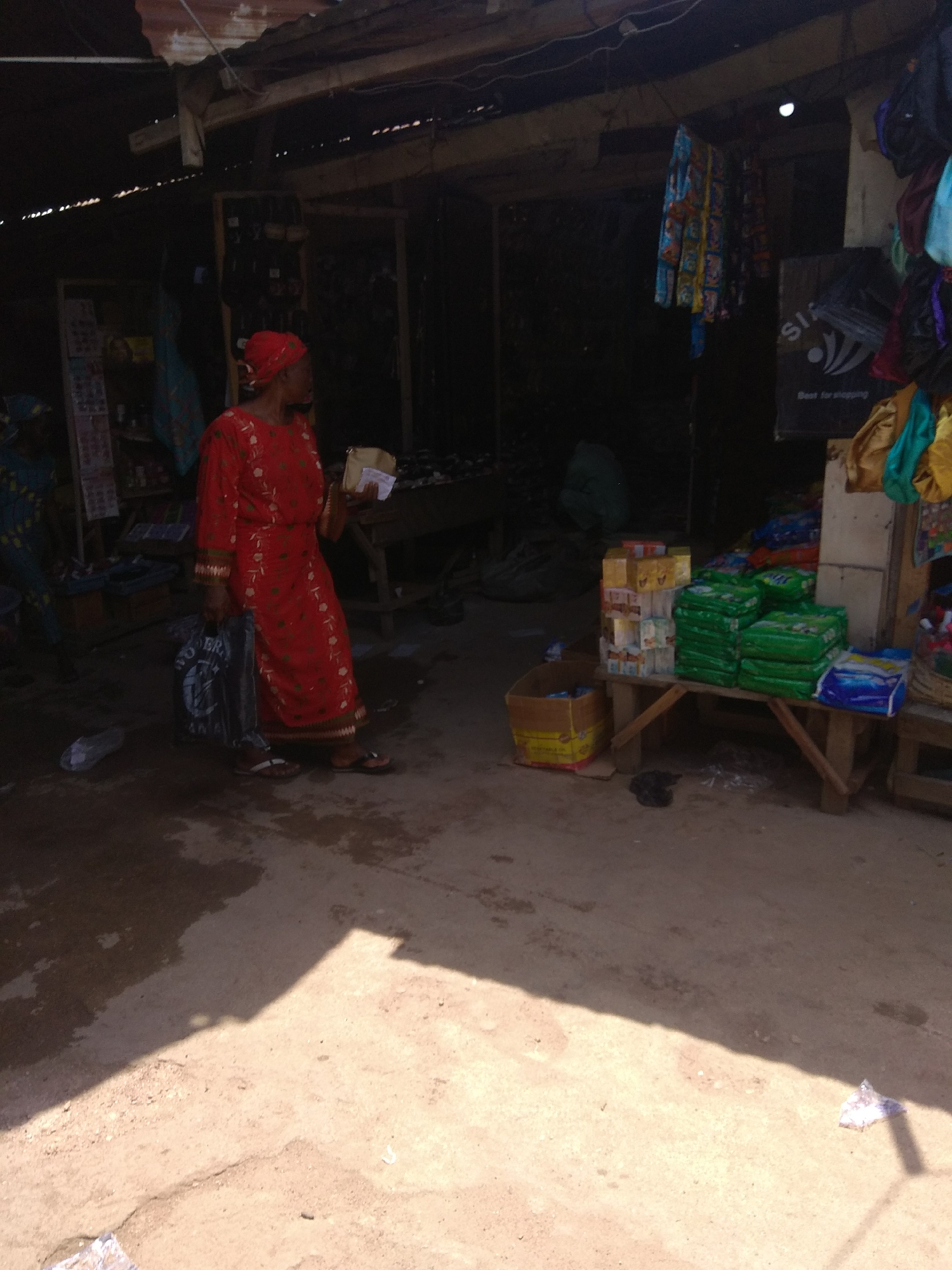 In Jalingo, Nigeria, a woman in a red dress stands in an outdoor market stall. She carries a blue bag in her left hand and a white paper in her right. The stall is filled with goods, including boxes of products stacked on a wooden bench and hanging clothing items. The ground is dirt with some puddles. The scene suggests a typical day in a Nigerian marketplace, with goods displayed for sale and shoppers interacting with vendors.