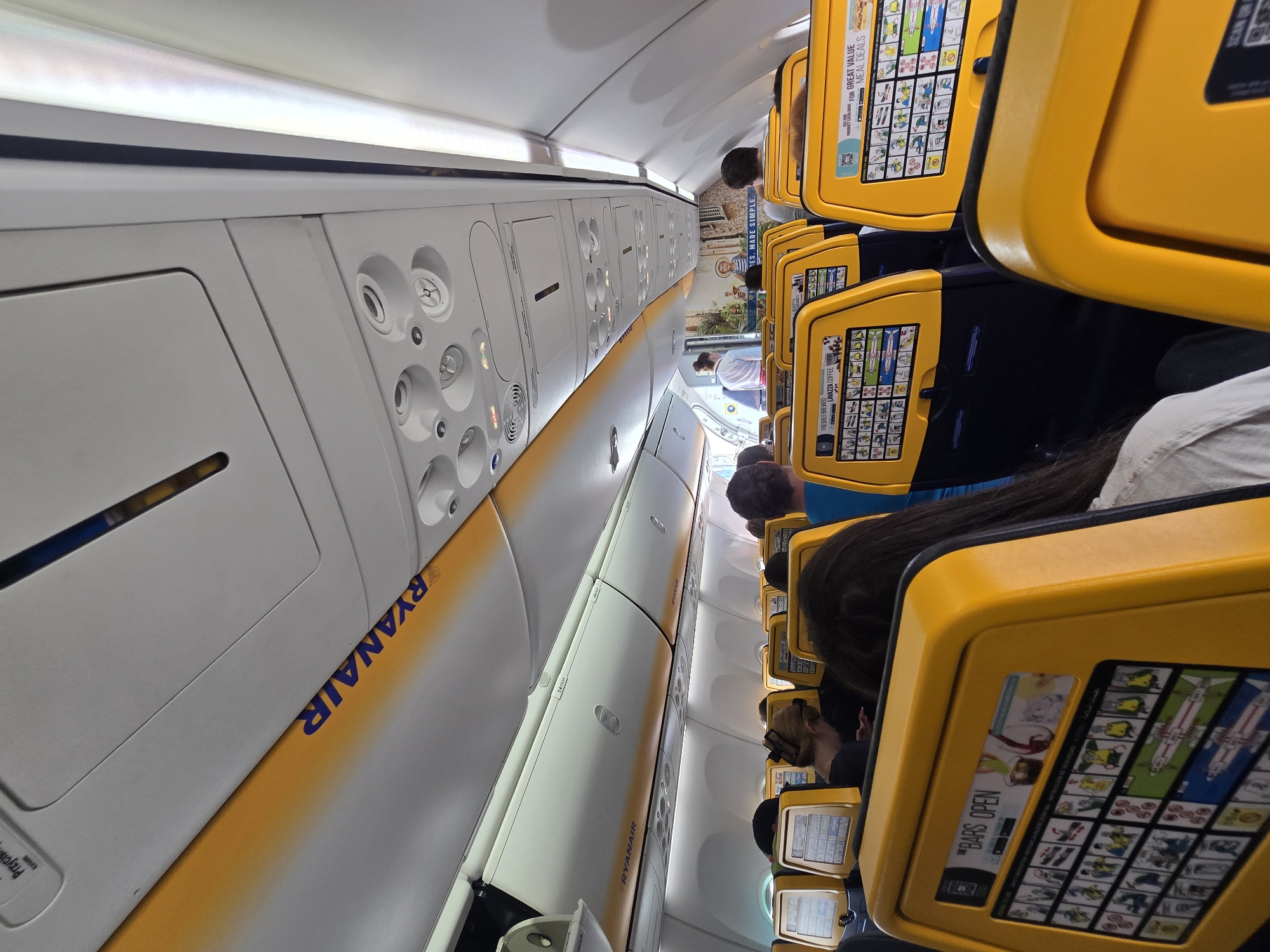 The image depicts the interior of an airplane cabin, likely a Ryanair flight, as indicated by the prominent yellow and blue branding and the airline's name visible on the overhead compartments. The perspective is from a passenger's seat, looking forward down the aisle. The seats are upholstered in dark blue fabric with yellow headrest covers, which also feature display screens. These screens display safety instructions, advertisements, and potentially entertainment options. In the foreground, a passenger's dark hair is visible, and to their left, a person with blonde hair tied up is looking at their phone. Further down the aisle, other passengers are seated, some looking forward, others partially obscured. Overhead, the typical airplane cabin features are visible, including air vents, lights, and storage compartments. A sign that says "RYANAIR" in blue letters is visible on the yellow trim of the overhead compartment. The lighting is artificial, typical of an airplane cabin, and the overall environment suggests a flight in progress.