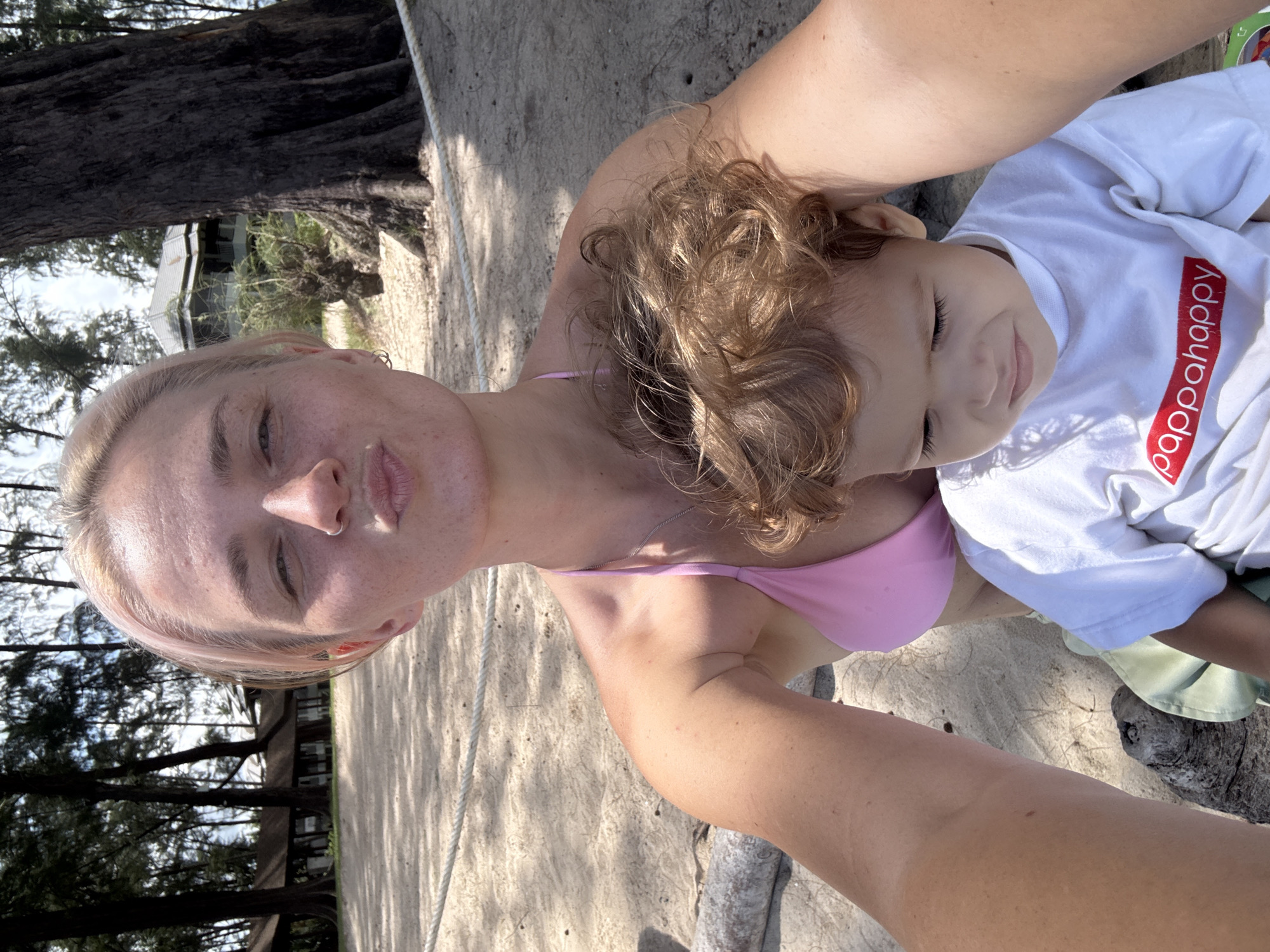 The media file shows a close-up selfie of two individuals, likely a mother and child, lying on a sandy beach. The woman in the foreground, with blonde hair and light skin, is wearing a pink bikini top. She is looking directly at the camera with a relaxed expression. Beside her, a young child with light brown curly hair is nestled against her. The child is wearing a white t-shirt with red lettering. The background features sand, tree trunks, and greenery, suggesting an outdoor, tropical setting. The lighting indicates it is daytime, with sunlight casting shadows on the sand, hinting at a sunny weather. The presence of trees and the beach setting strongly suggest a location like Choeng Thale, Thailand. The overall mood appears to be one of closeness and relaxation. The visible text on the child's t-shirt reads "pappahappy" in red.