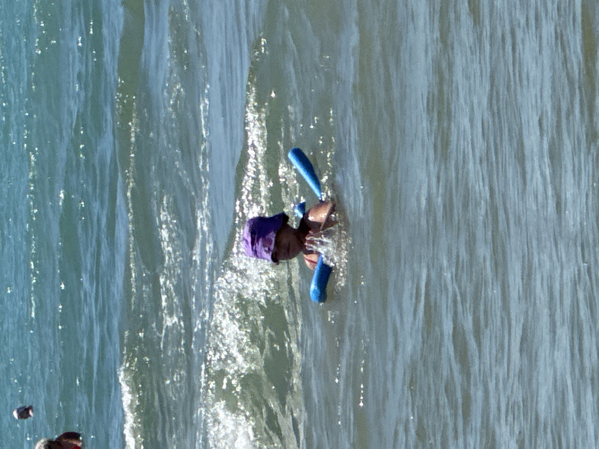 A young child wearing a purple bucket hat and a pink swimsuit is in the water, holding a blue pool noodle. The child is positioned in the middle of the frame, with waves around them. The water appears to be a bright blue with sunlight glinting off the surface. In the background, there are hints of other people in the water, and some blurry landmass in the far distance. The scene is set in a body of water, likely the sea or a large lake, with moderate waves.