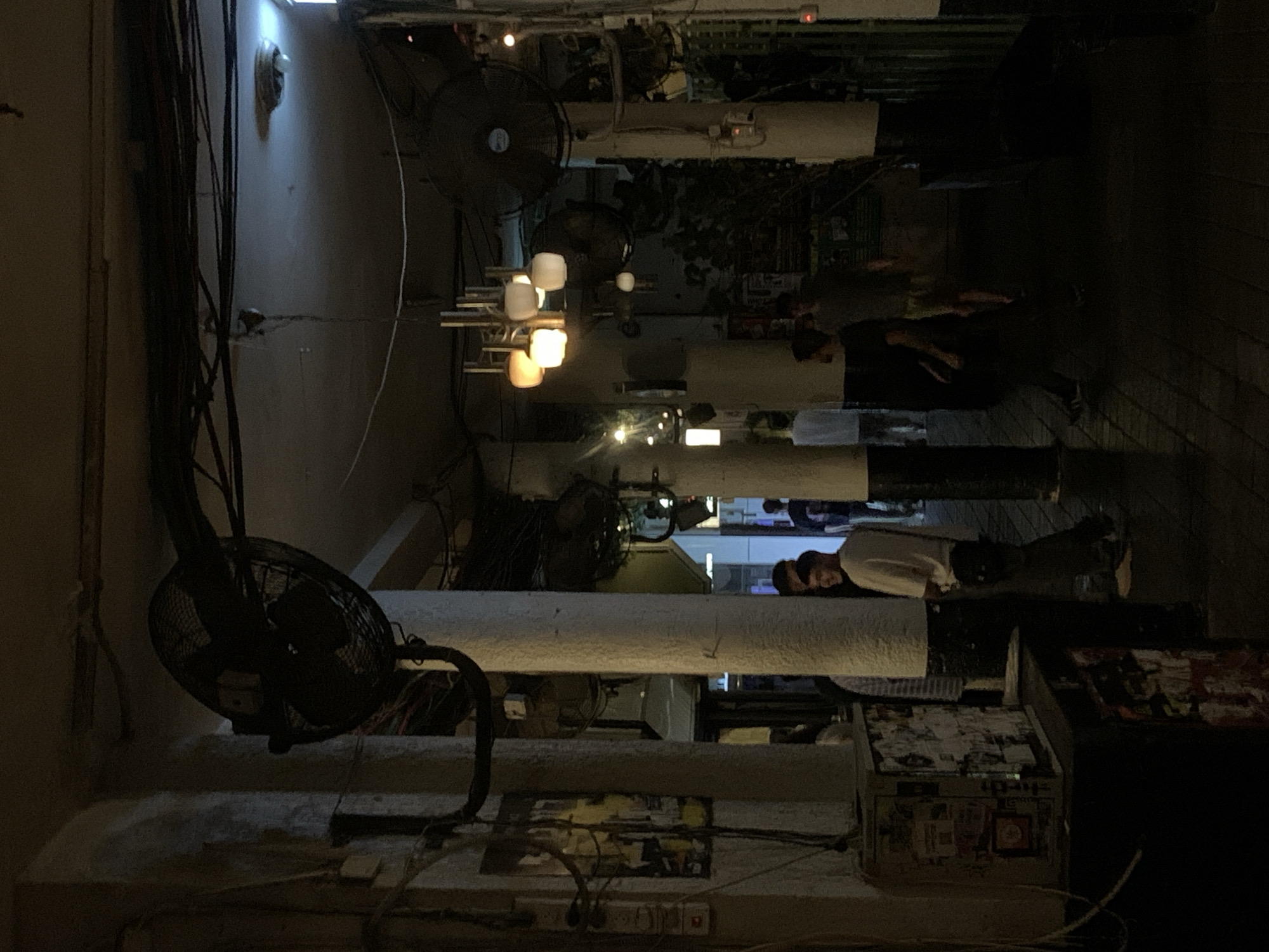 This image captures a dimly lit, narrow, urban passageway located in Tel-Aviv, Israel, during the evening or night. The environment is characterized by an aged, utilitarian aesthetic with exposed infrastructure.
The scene is structured by multiple thick, round, light-colored columns that line both sides of the passageway. The bases of some columns are painted dark. A dense network of black electrical wires, cables, and conduits runs haphazardly along the walls and across the low ceiling. At least three large, dark-colored industrial wall-mounted fans are visible, two on the left side (one prominent in the upper left foreground, another in the mid-ground left) and one on the right side in the mid-ground.
Illumination is provided by various artificial sources: a central multi-bulb chandelier with warm-toned, opaque glass shades hangs from the ceiling in the mid-ground. Additional localized lighting comes from exposed bulbs and strip lights further down the passageway, revealing glimpses of adjacent spaces or shopfronts. Electrical boxes and switches are mounted on some columns. On the lower left, a utilitarian cabinet or box is heavily covered with an array of stickers and advertisements, adding to a cluttered, urban texture. The floor is paved with dark-colored tiles or pavers.
Approximately seven to ten individuals are present in the passageway. In the mid-ground, a young adult male wearing a white t-shirt and dark pants, with a small black bag slung across his body, is seen smiling and looking towards the right, suggesting social interaction. He stands near a column. Another individual in a striped shirt is partially visible behind a column on the left. Further down the passageway, other figures are observed walking or standing; one person in a light-colored shirt is seen from behind, walking away, and another person in a dark t-shirt and shorts is visible further right, moving left. The individuals are casually dressed. The overall activity indicates a casual social gathering or movement through a commercial or entertainment thoroughfare.