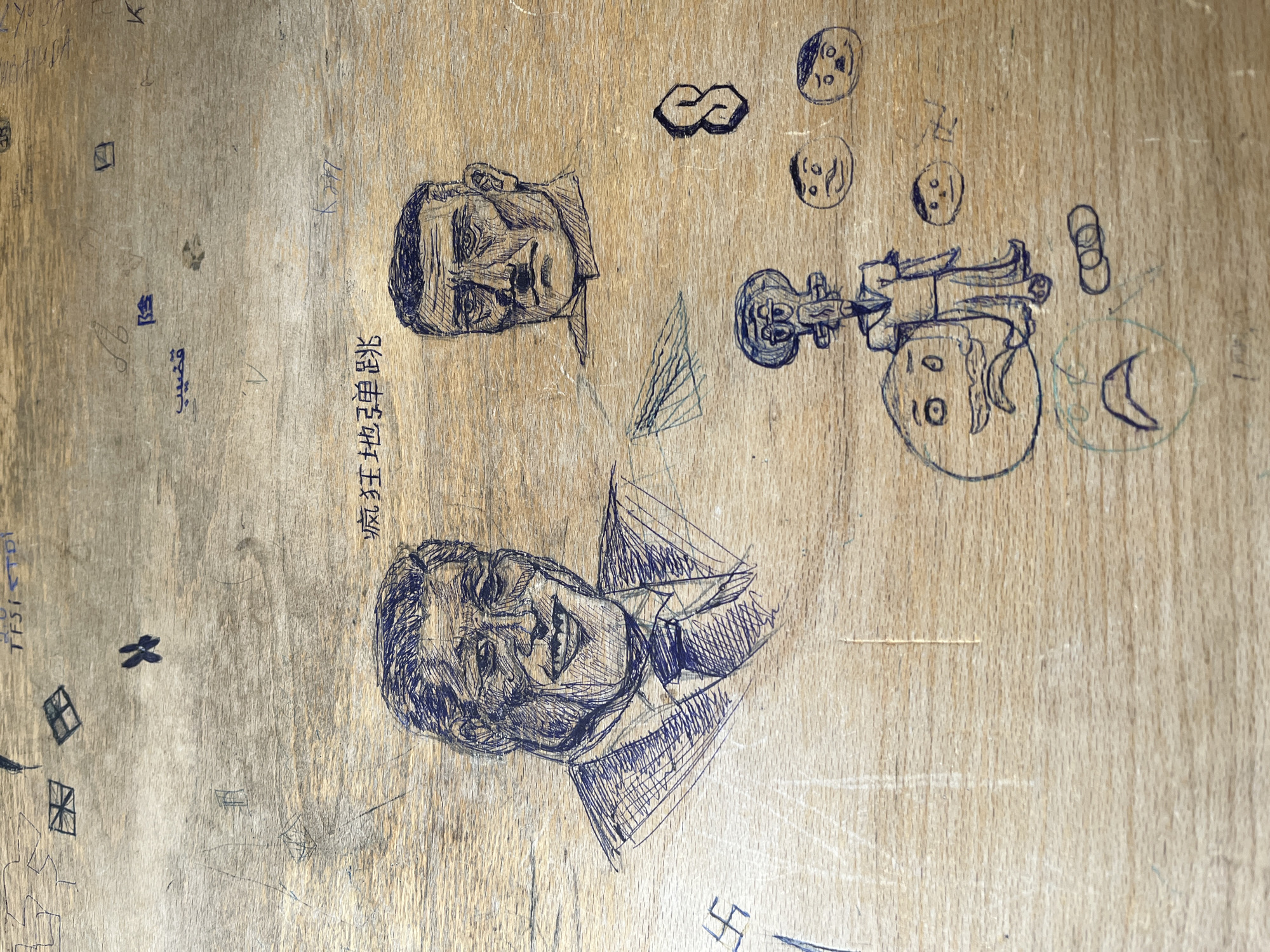 The image displays a close-up of a light-colored wooden surface, possibly a desk or panel, covered with various pen drawings and scribbles. The overall impression is that of spontaneous graffiti or doodles.
The main subjects are two detailed caricatures of men, drawn in blue ink. One, positioned in the upper center, depicts a man with a neutral or slightly serious expression, broad forehead, and short hair. The other, located below it, shows a smiling man with a prominent jawline and short hair, wearing a collared shirt and tie, with cross-hatching used for shading.
Between these two caricatures, Chinese characters are written vertically: "张国荣拜王" (Zhāng Guóróng bài Wáng), which translates to "Leslie Cheung worships Wang."
Other doodles scattered across the surface include:
- An infinity symbol and several simple circular faces with varying expressions in the upper right.
- A figure resembling Squidward from Spongebob Squarepants, next to a circular head with wide eyes and a smile, and a crescent moon.
- Various abstract shapes, such as triangles, rectangles, and concentric circles.
- A small butterfly-like symbol and a distinct swastika symbol towards the bottom right.
On the left side of the surface, there are faded and partially legible scribbles:
- Some illegible Arabic-like script.
- English-like text that reads "TFSI TD!", possibly referring to car engine designations.
- Other faint letters and numbers, including a "K" and small square outlines.
The wooden surface itself shows natural grain, minor scratches, and wear, indicating its age and use as a canvas for these drawings. The drawings are oriented such that they appear mostly upright, suggesting the photo was taken from a specific angle or the original surface was positioned vertically.