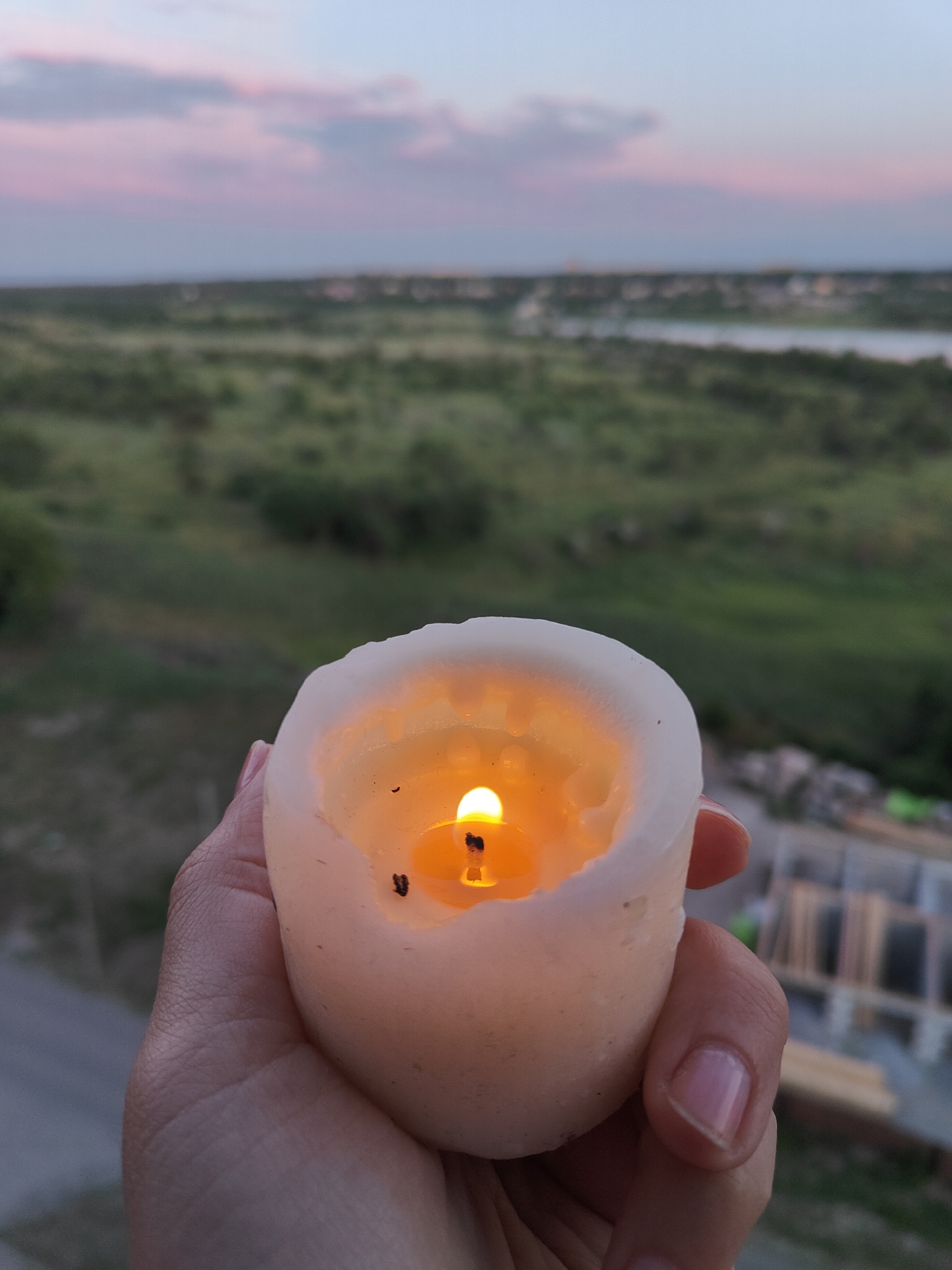 A hand holds a lit white candle. The candle is partially melted, with a pool of wax visible around the flame. The background shows a blurred landscape with green fields, a body of water, and a hazy horizon with buildings. The sky above is a soft gradient of pale blue and pink, suggesting twilight or dawn. The overall scene appears to be from the outskirts of Kryvyi Rih, Ukraine, possibly overlooking an industrial or developing area given the visible construction materials in the far background.