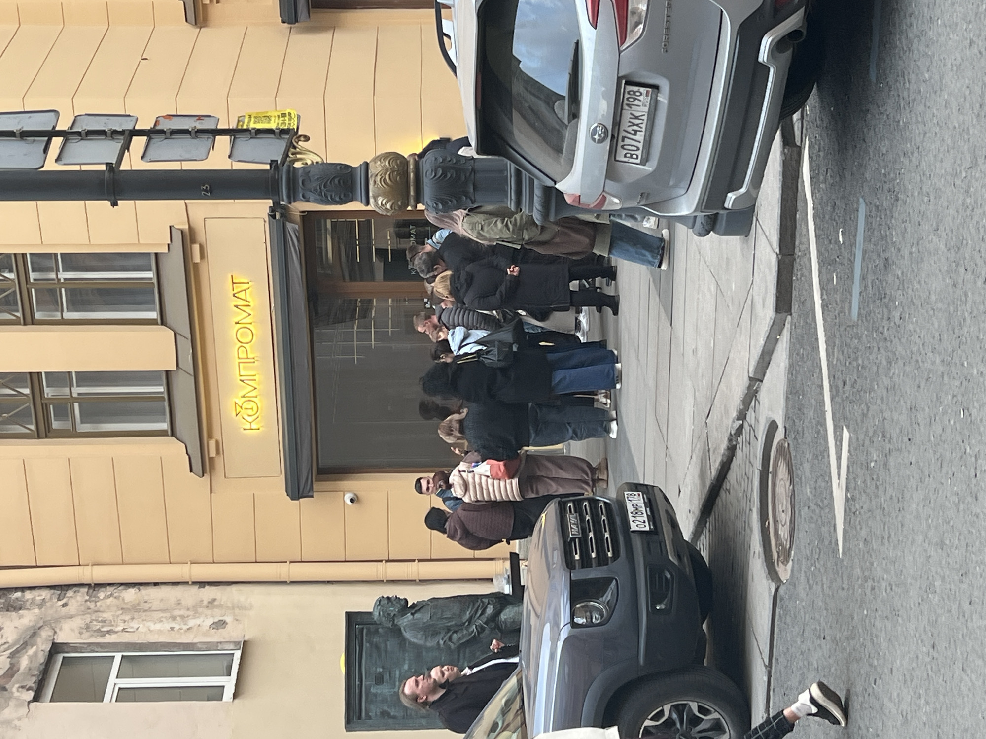 This daytime image, captured in Saint Petersburg, Russia, depicts an urban street scene with a notable queue of people. Approximately 15-20 individuals are lined up on the sidewalk in front of a light-colored building that features a prominent, glowing yellow neon sign displaying the Cyrillic text "КОМПРОМАТ" (KOMPROMAT). The people in the queue are dressed in various casual outfits, including jackets and coats, suggesting cool but clear weather.
On the street alongside the sidewalk, two vehicles are visible. A silver Subaru Forester is parked or stopped, its rear showing the Russian license plate B074XX 198, with '198' indicating the Saint Petersburg region. Further down, the front of a dark SUV, possibly a Haval, is visible with the license plate 0218HP 178, '178' also being a Saint Petersburg region code. The asphalt road surface includes painted white lines and a circular manhole cover.
A black street lamp pole with decorative gold accents stands near the building's entrance, bearing the number "23." Partially obscured by the dark SUV and parked cars, a dark-colored statue of a seated figure is visible on the sidewalk. The building itself is a light beige or cream color with multiple windows. The scene conveys a bustling urban environment, possibly around a popular establishment or event that has drawn a queue of people.