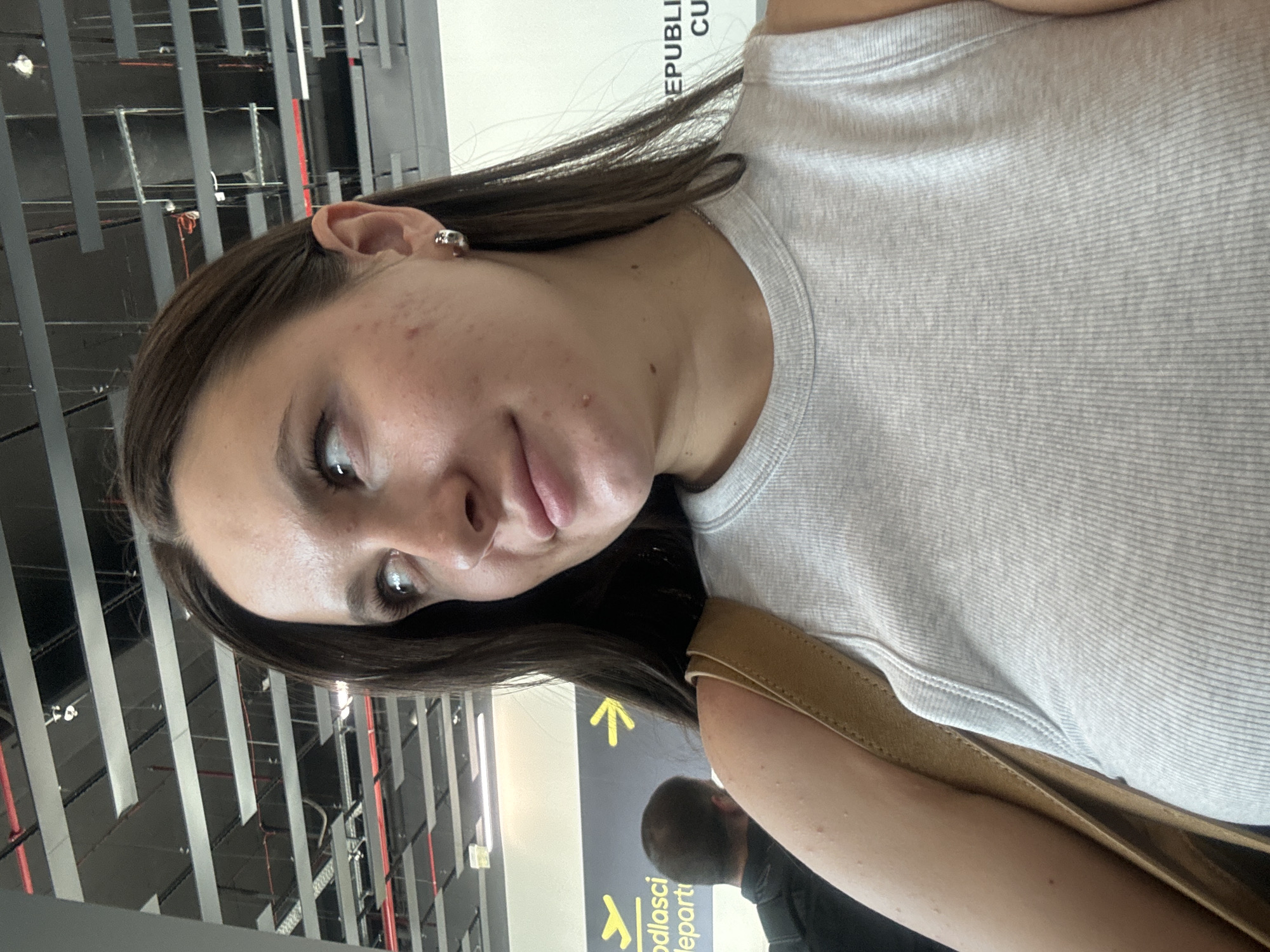 A woman with dark hair is at Belgrade Nikola Tesla Airport in Surcin Urban Municipality, Serbia. She is wearing a light gray ribbed tank top and a brown bag strap is visible on her shoulder. She has an earring in her left ear. In the background, there is a sign in Serbian and English for "departures," with a yellow arrow. Behind her is a man with dark hair and a dark shirt. The airport's ceiling structure is visible.