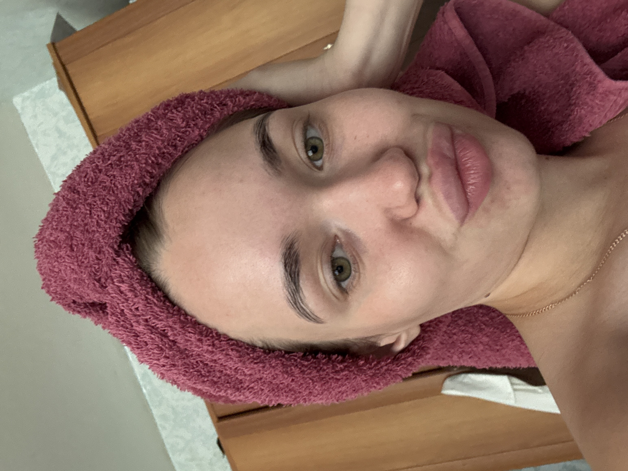 A close-up, rotated, eye-level shot captures a fair-skinned young woman lying on what appears to be a wooden surface, possibly a massage table or a bed. A pink or maroon-colored towel is wrapped around her head, framing her face. Her eyes are open, and she appears to be looking directly at the camera. She has prominent eyebrows and full lips. Part of a thin, gold chain necklace is visible on her neck. The background is slightly blurred, with hints of a white surface and the wooden table. The location and city of Chaikovsky, Russia, cannot be verified from the image alone.