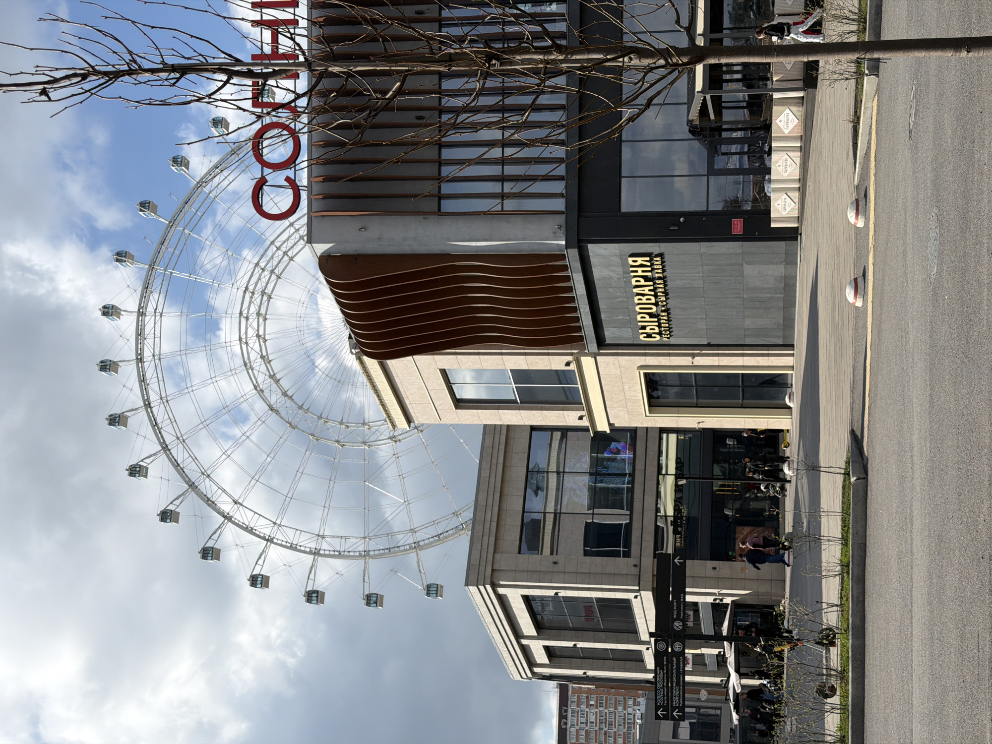 This media file captures an urban scene in Moscow, Russia, featuring the "Sun of Moscow" Ferris wheel prominently on the left. The large, white metal structure of the Ferris wheel, with its numerous enclosed cabins, dominates the left half of the image, set against a partly cloudy sky.
In the foreground and mid-ground, several modern multi-story buildings line a paved street and sidewalk. The architecture is contemporary, characterized by large glass windows, clean lines, and some decorative elements like horizontal wooden slats on one building and wavy, patterned panels below it.
On one of the buildings, a prominent sign displays the text "СЫРОВАРНЯ" (Syrovarnya), with smaller text beneath reading "РЕСТОРАН • СЫРНАЯ ЛАВКА" (Restoran • Syrnaya Lavka), indicating a restaurant and cheese shop. Above this building, partially visible red letters on another structure spell out "СОЛНЦЕ..." (Solntse...), likely part of "Солнце Москвы" (Sun of Moscow), referring to the Ferris wheel itself.
The street is asphalt, bordered by a concrete sidewalk. Several small, bare-branched trees are scattered along the sidewalk, suggesting late autumn, winter, or early spring. A few people are visible on the sidewalk; some appear to be walking, while others are seated at outdoor tables under umbrellas, indicating a cafe or restaurant area. White and red striped bollards are placed along the edge of the street.
The overall scene depicts a lively, modern urban area, likely a commercial or entertainment district, with the iconic Ferris wheel serving as a major focal point. The sky is bright but mostly overcast.