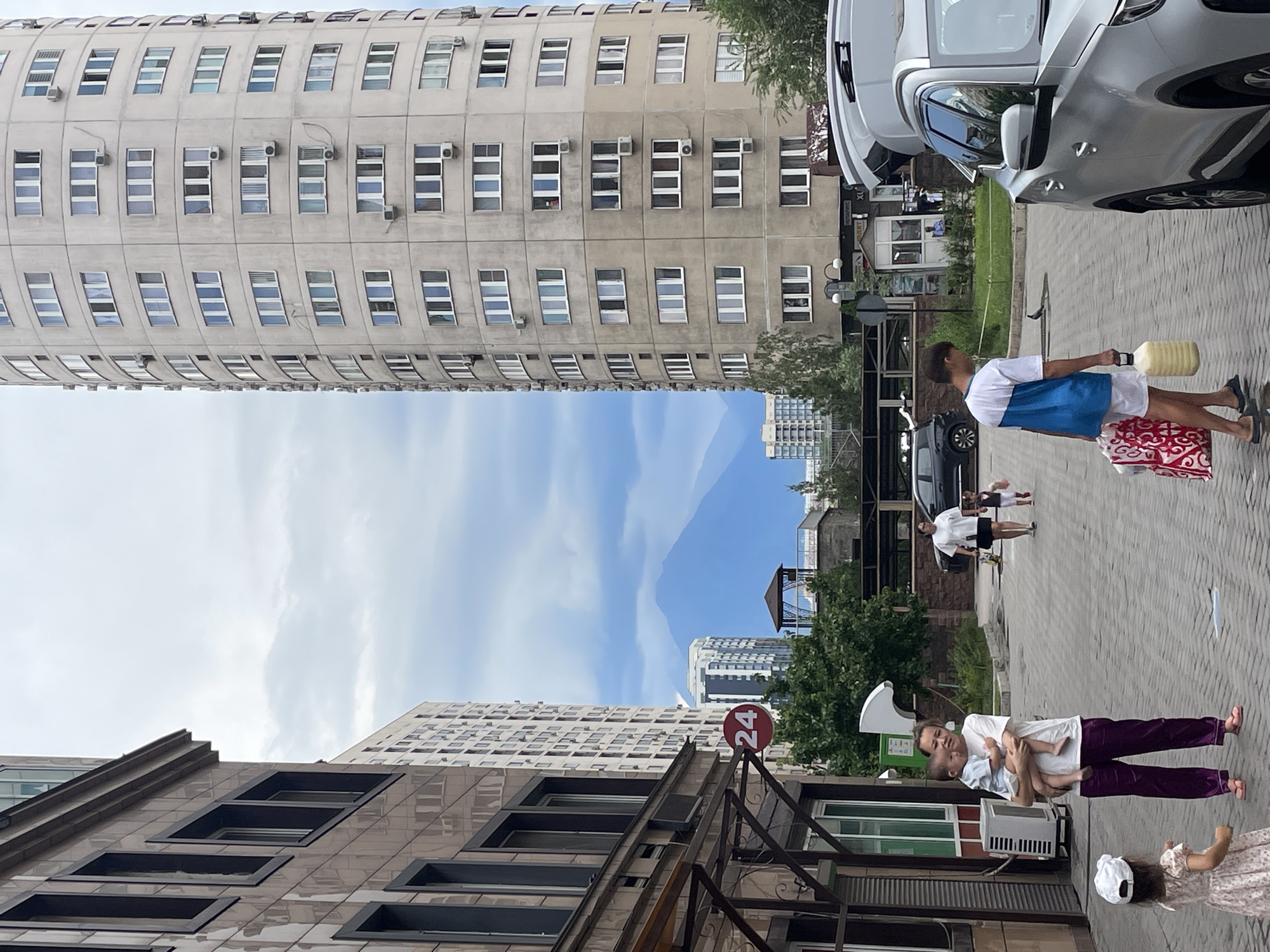 In Almaty, Kazakhstan, on a paved city street, a mother holds a baby while standing next to a building with a dark tiled facade. Across the street, a tall, cylindrical building with numerous windows and air conditioning units dominates the skyline, with mountains visible in the background under a cloudy sky. A young girl in a dress and hat stands to the left of the mother. Further down the street, a young man carries a large yellow container and shopping bags towards a silver SUV. Other people and a black car are also present. A red circular sign with the number "24" is visible on a pole.
