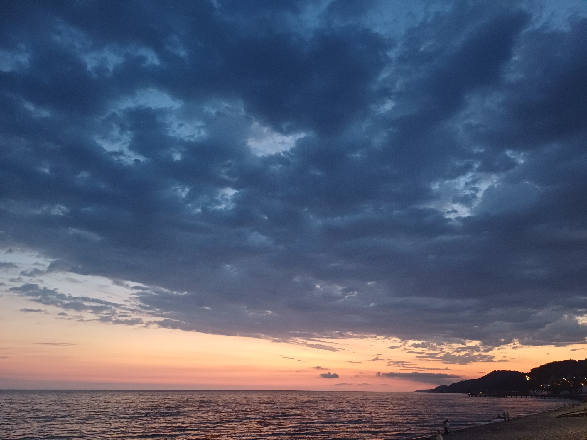 The image depicts a twilight scene on a beach in Sochi, Russia, likely during the evening. The sky is dominated by dark, dramatic clouds, with a sliver of pastel orange and pink light visible near the horizon, indicating the setting sun. The sea is calm, with gentle waves lapping the shore. In the distance, hills and a small town with lights are visible along the coastline. A pier extends into the water, and a few figures are scattered on the sandy beach, some walking and others possibly seated. The overall atmosphere is serene and somewhat melancholic.